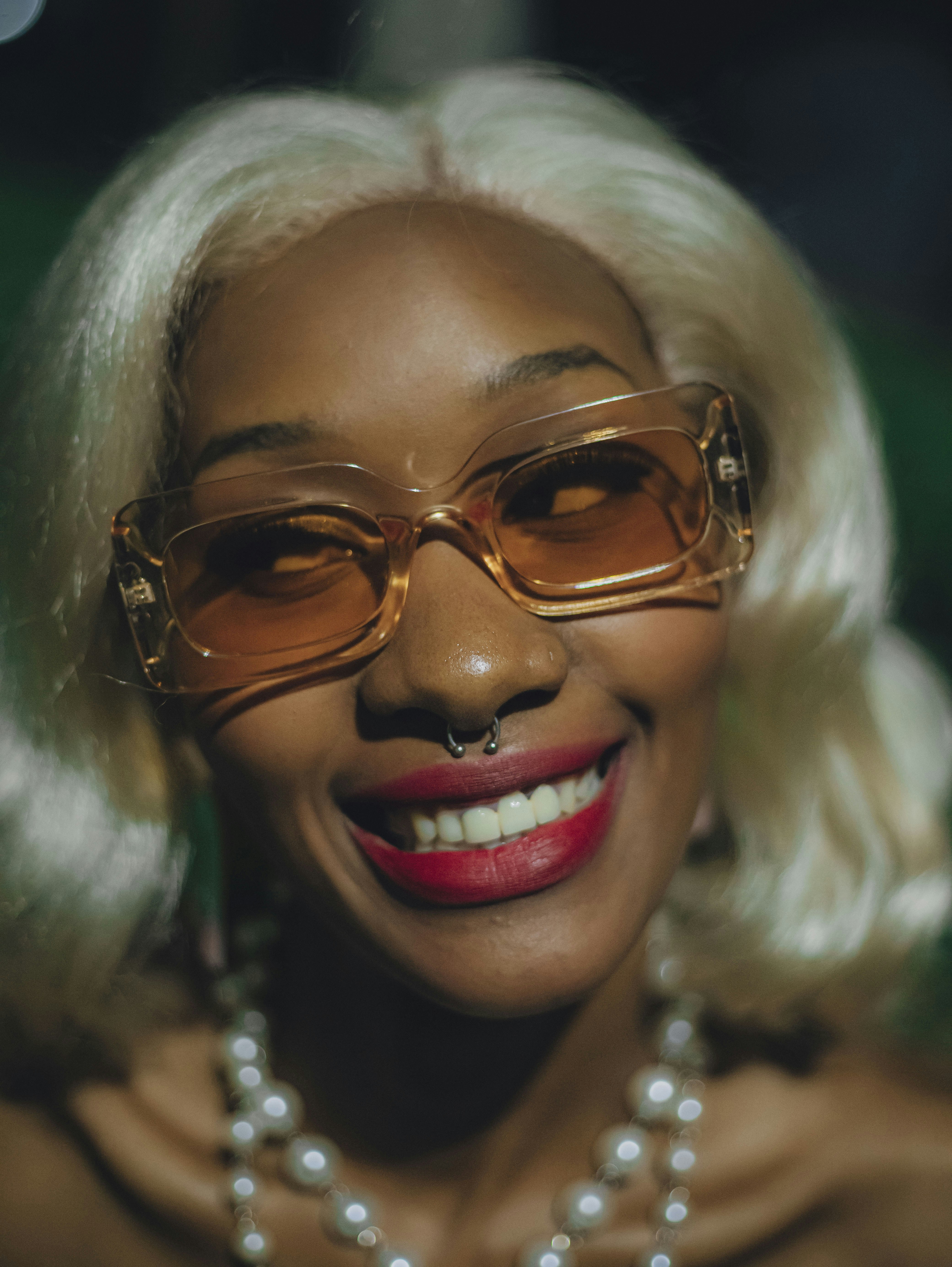 Mujer rubia sonriendo, con gafas de sol y un collar de perlas.