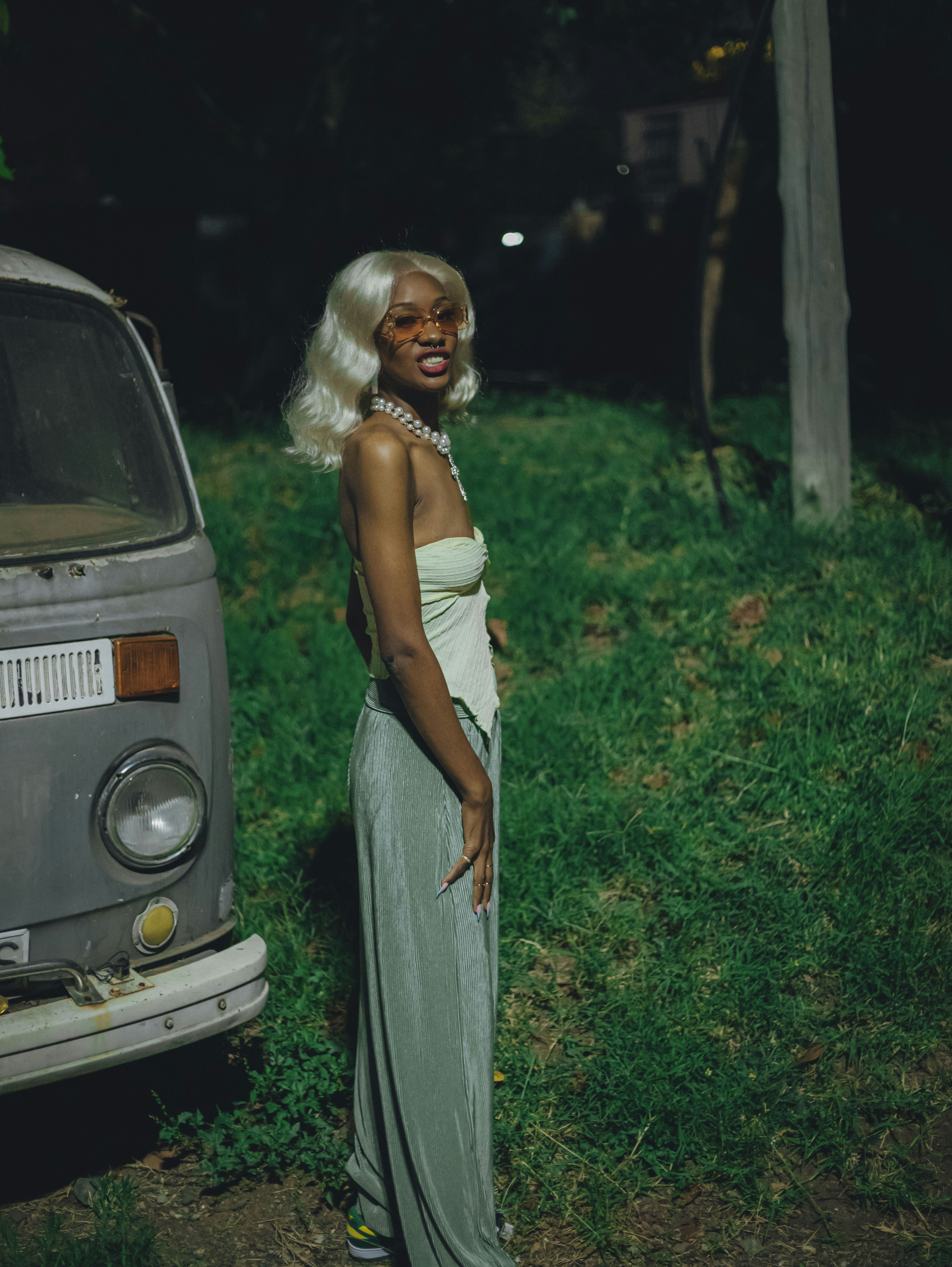 Mujer rubia vestida de verde de pie junto a la furgoneta vintage por la noche.