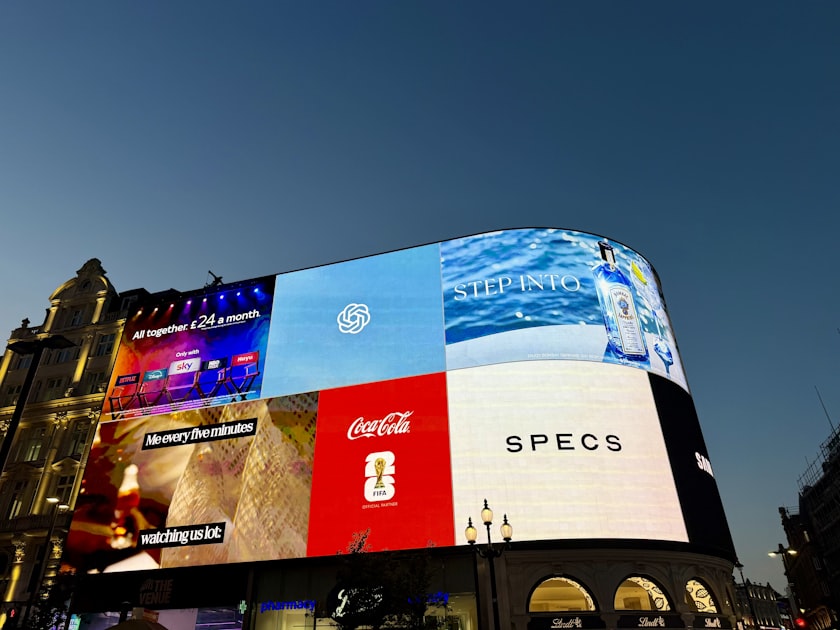 Large illuminated advertising billboards on the side of a building at dusk, representing the arrival of paid placement inside AI assistants