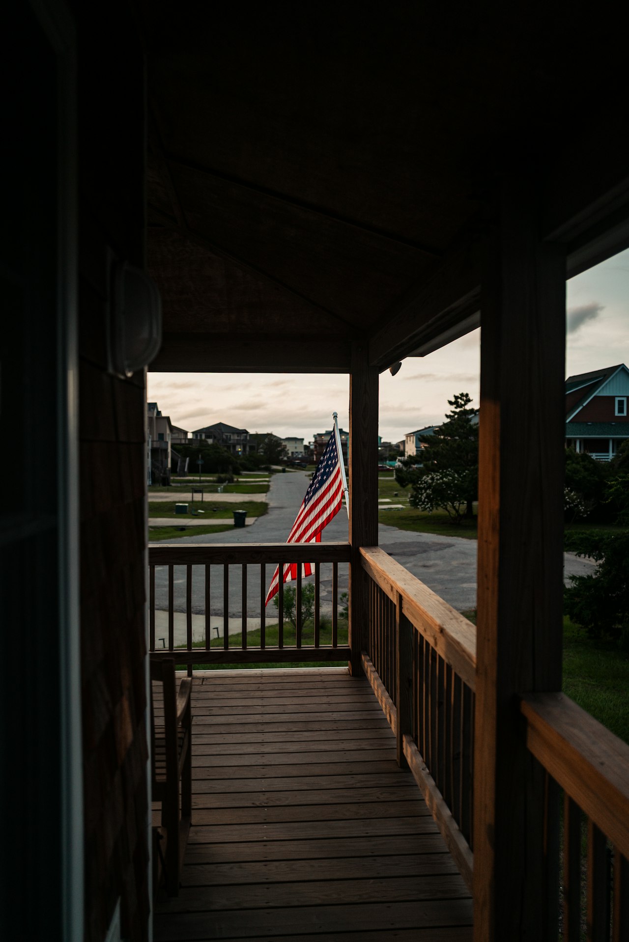 Covered wooden porch overlooking a residential street, with an American flag hanging beyond the railing.