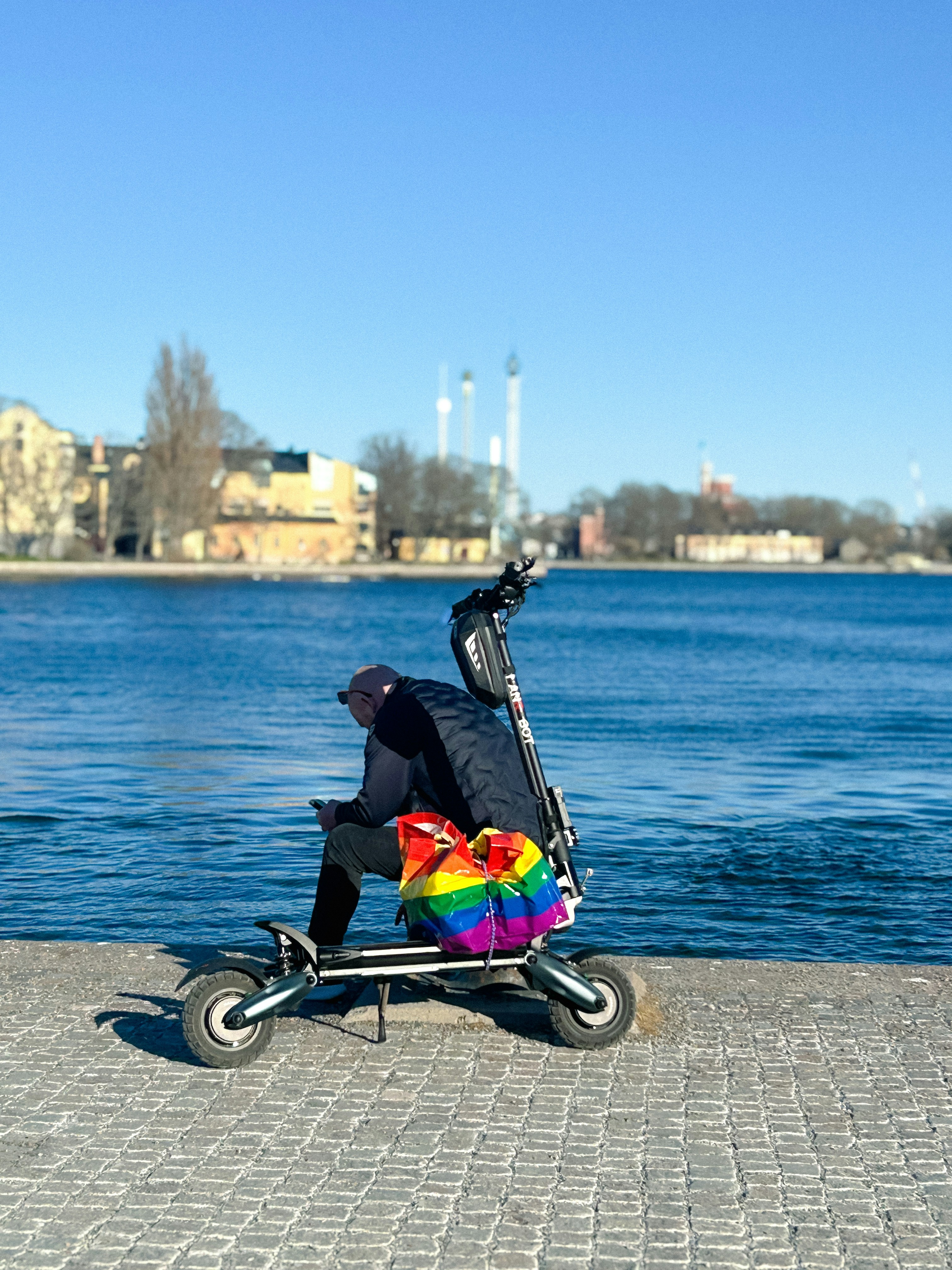 Person on scooter with rainbow bag by calm blue water.