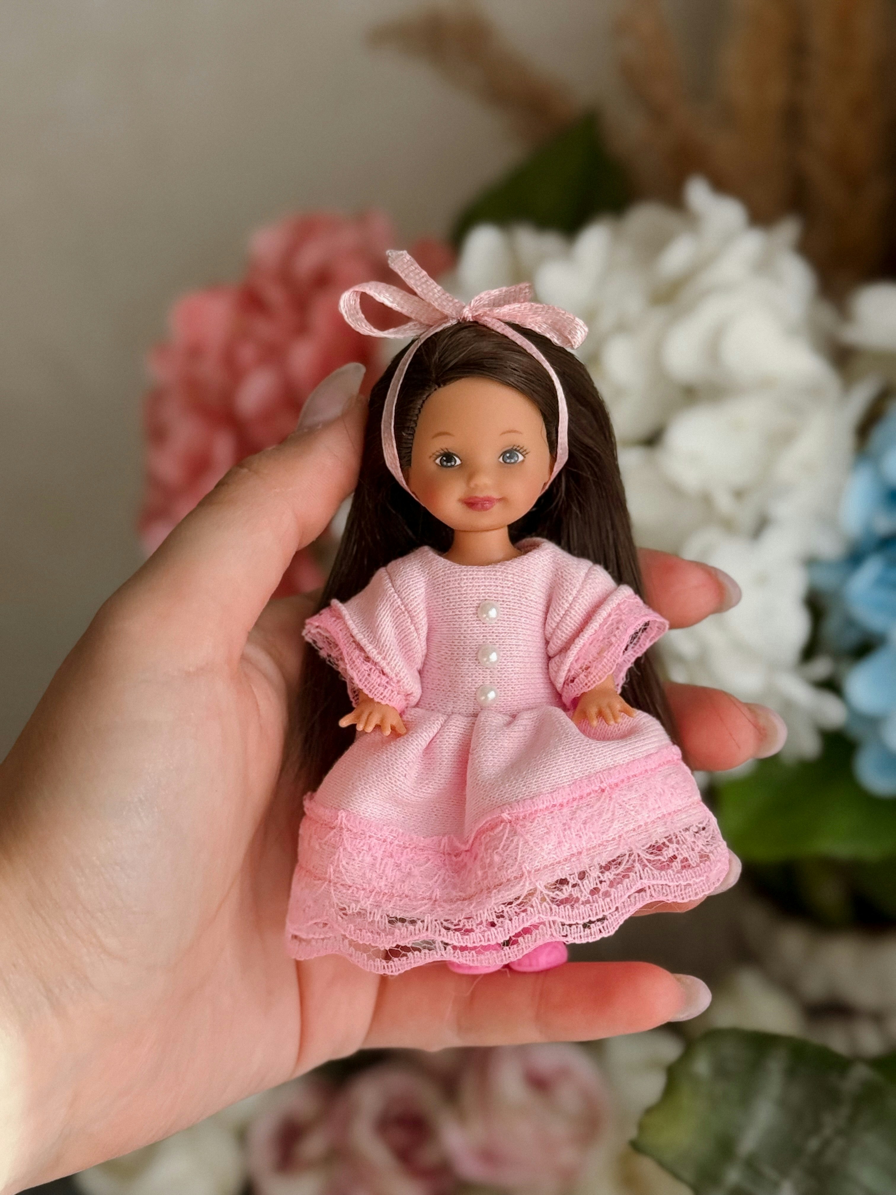 Une main tient une petite poupée vêtue d’une robe rose.
