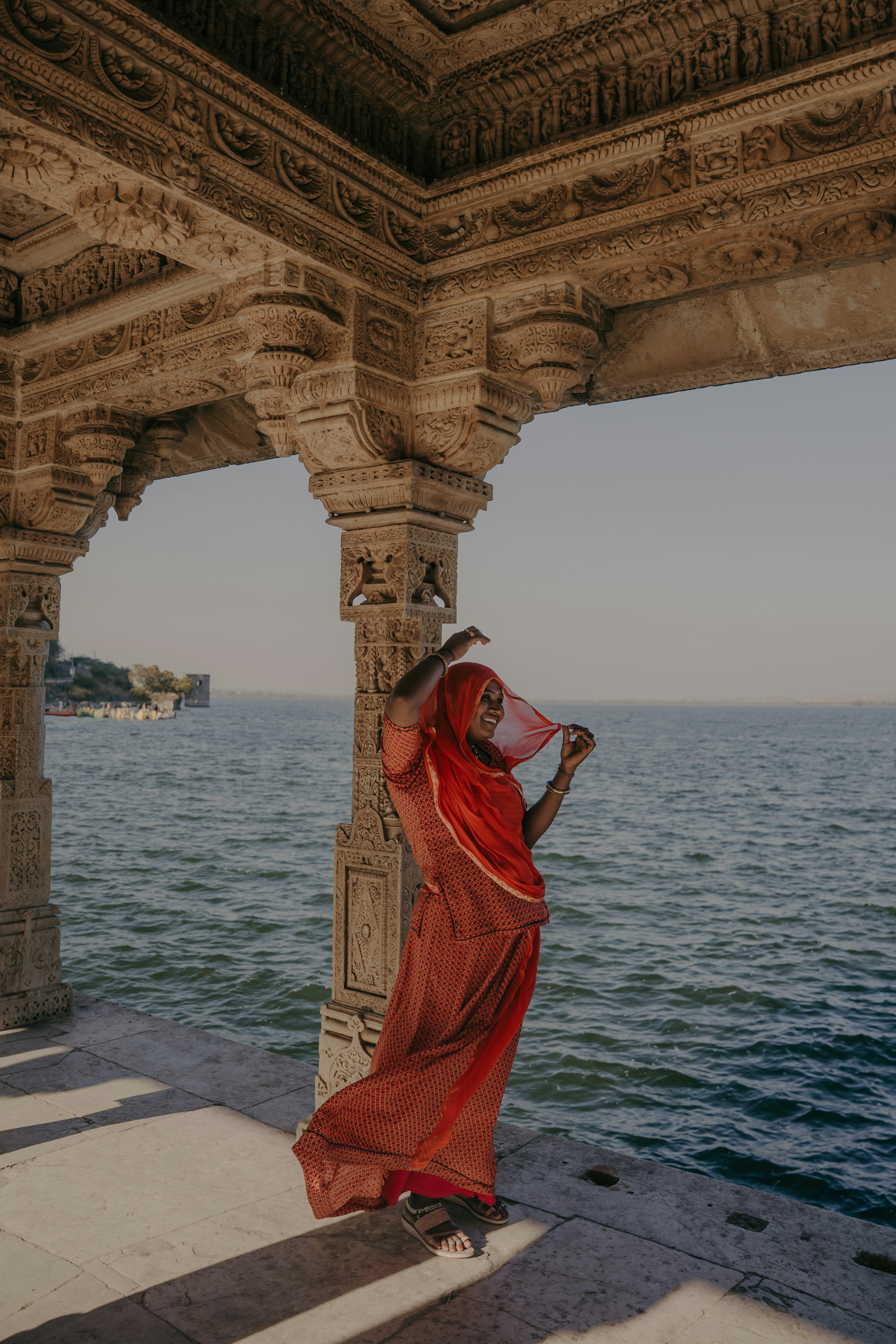 Woman in red sari under stone archway by a lake.
