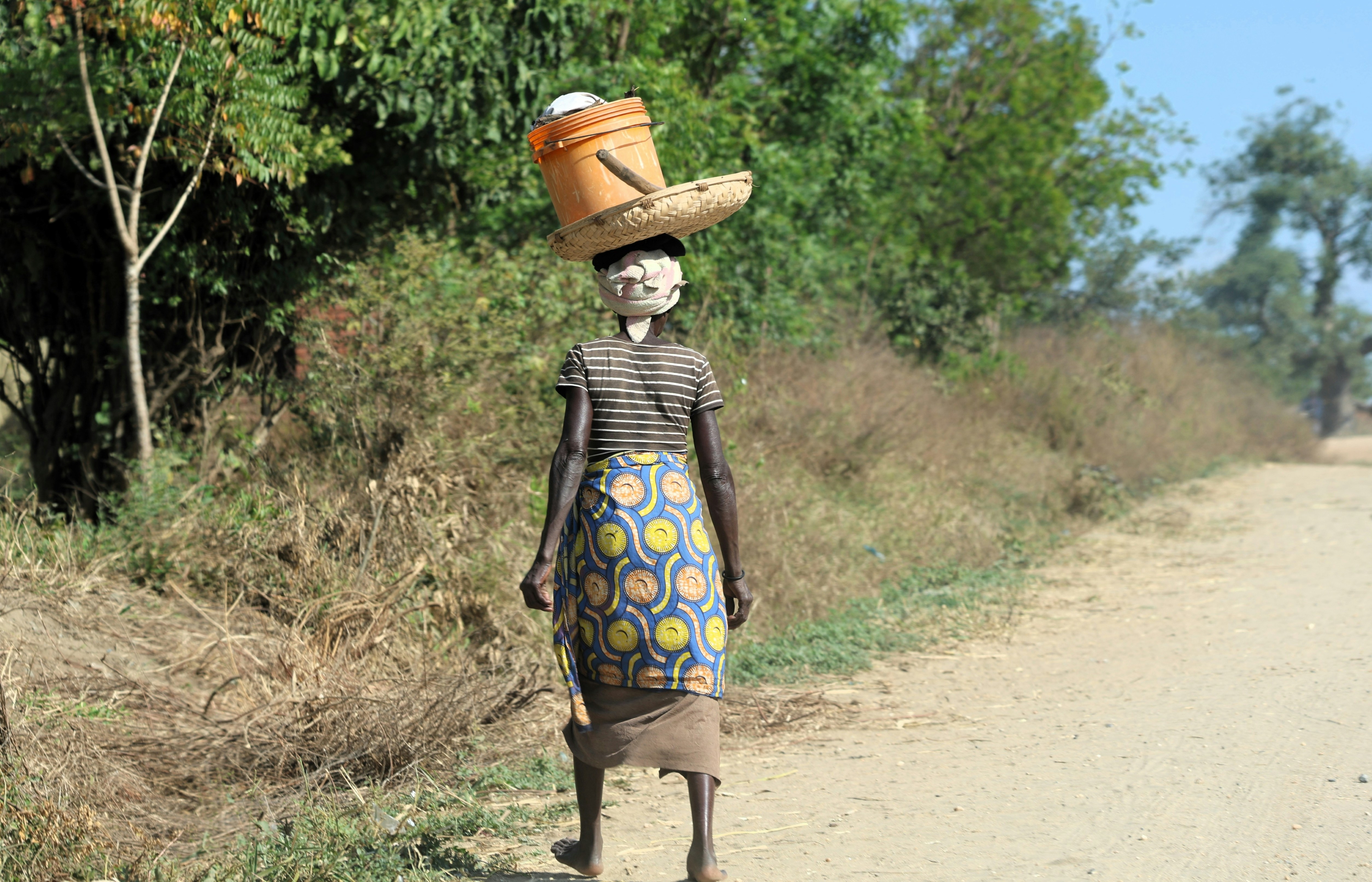 Woman carries bucket and hat on head along dirt road