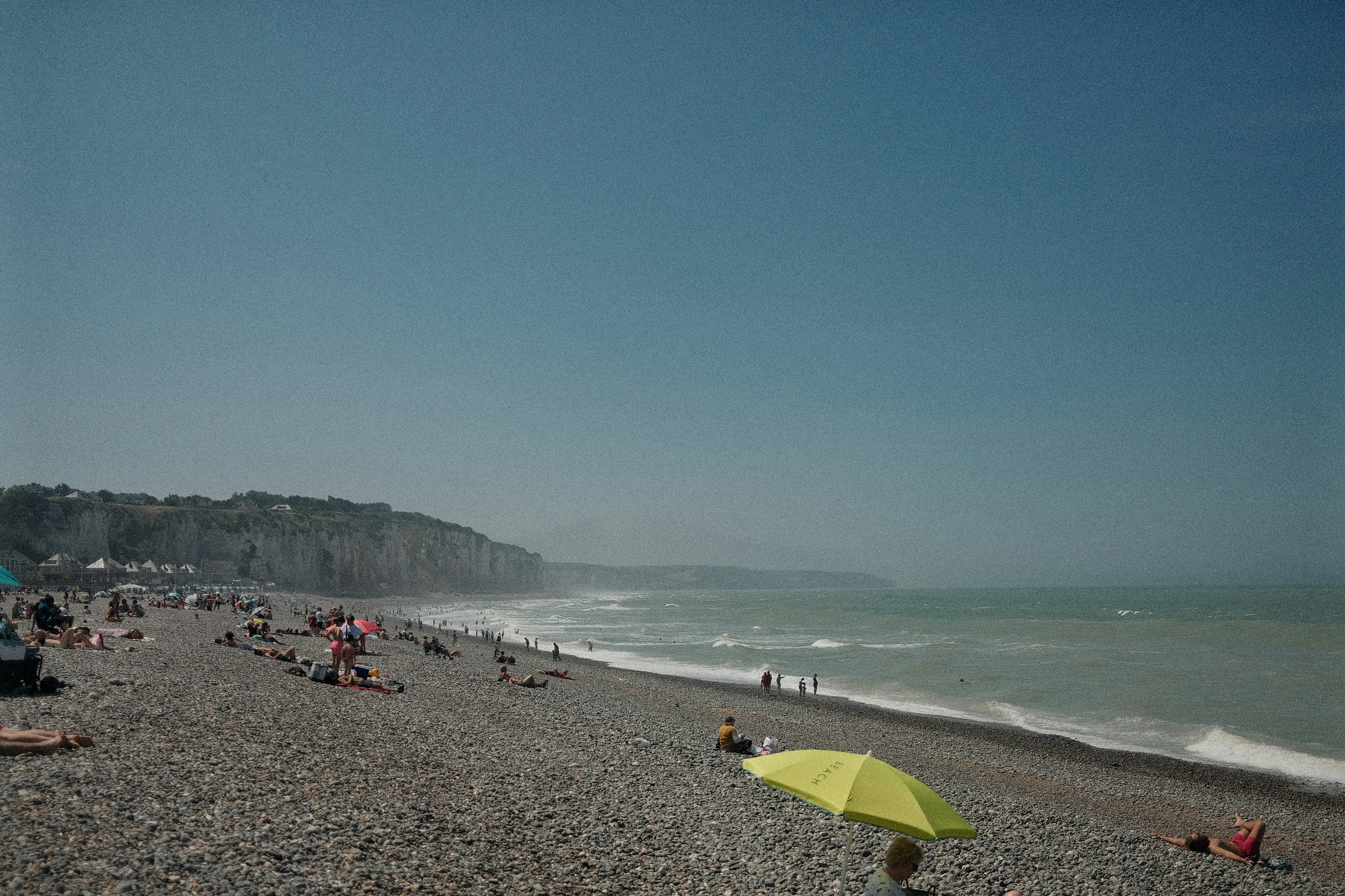 Sunny pebble beach with people, ocean waves, and distant cliffs.