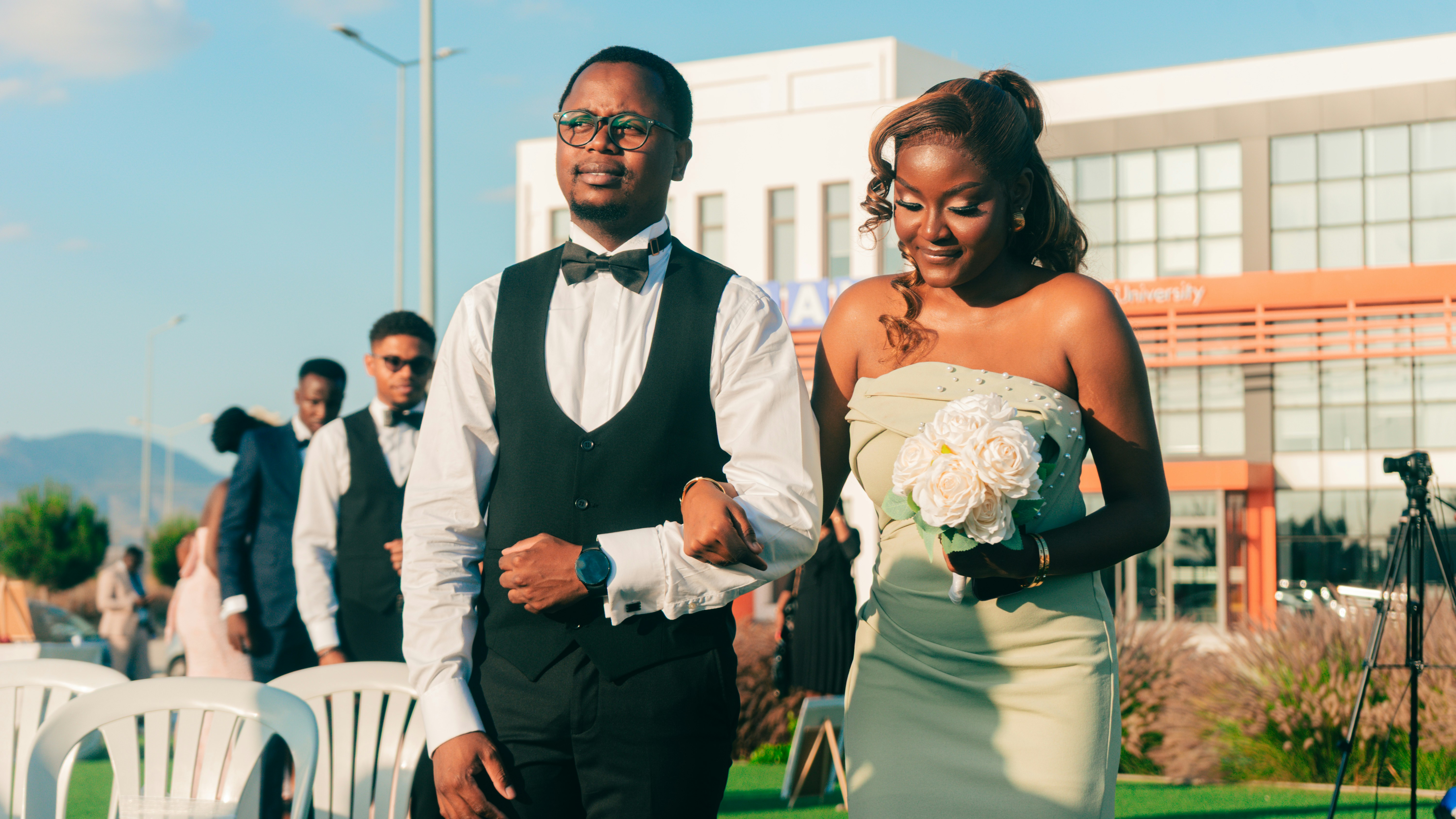 Man and woman in formal attire walk arm-in-arm with flowers.