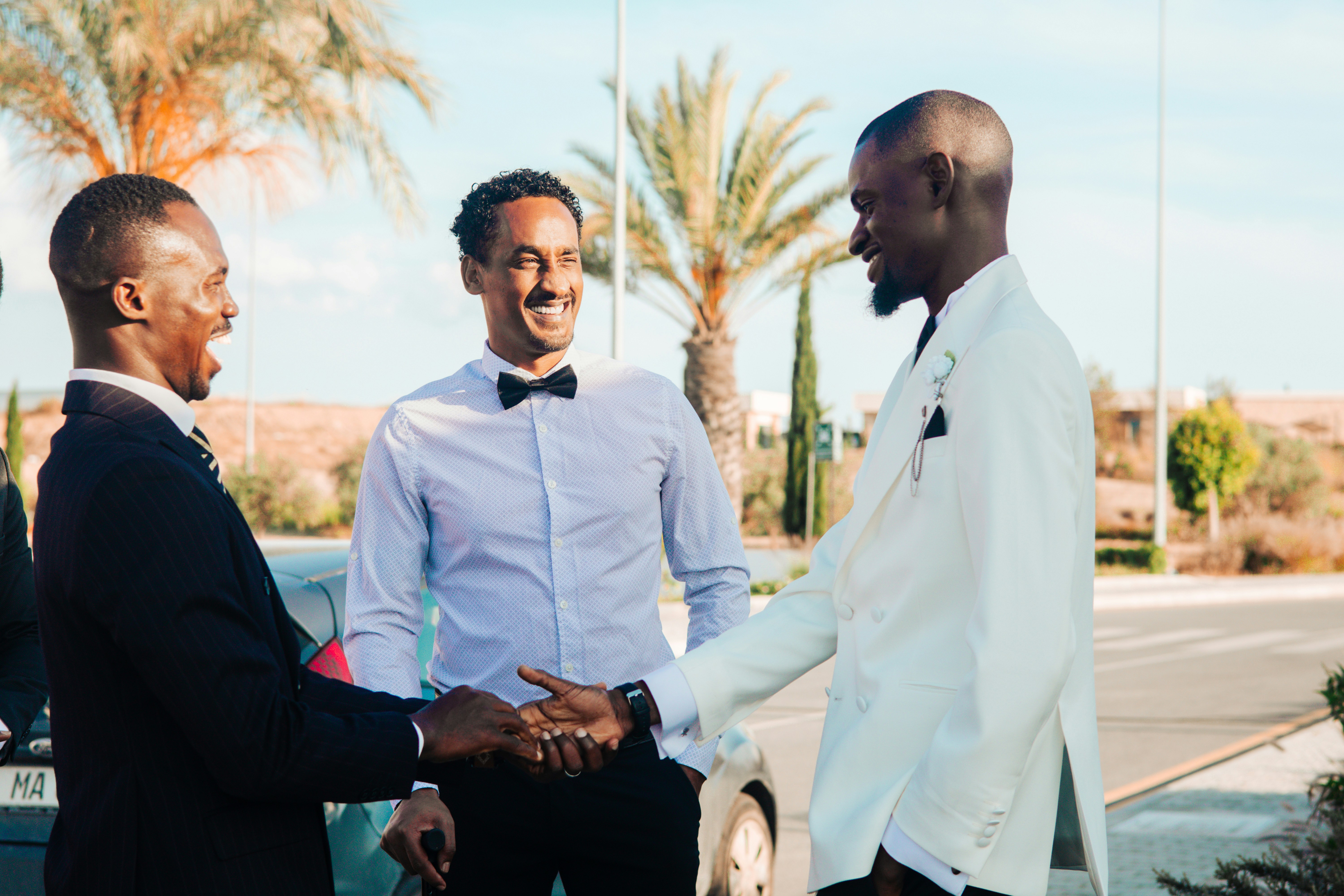 Three formally dressed men shaking hands and laughing outdoors.