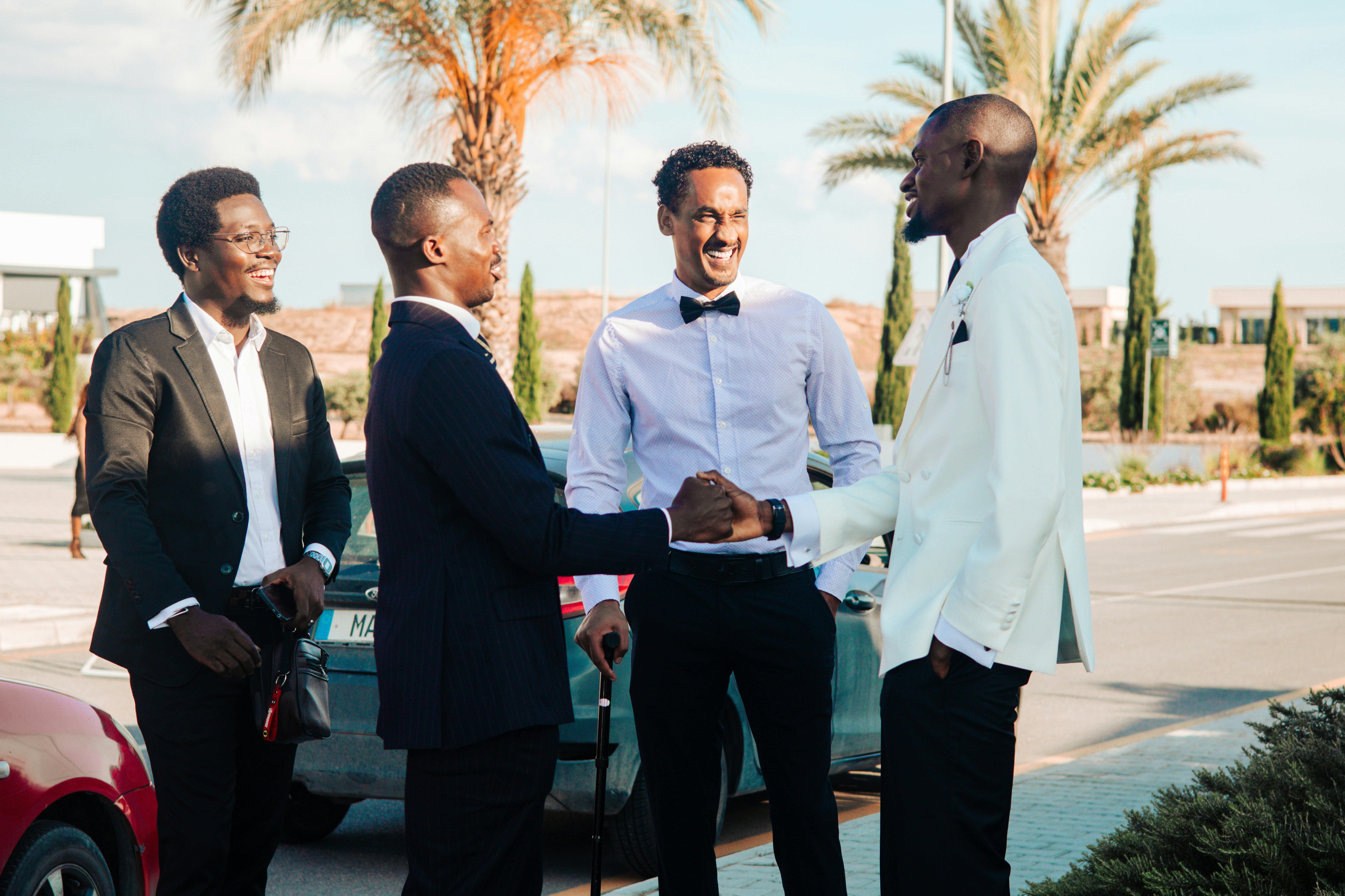Four formally dressed men shaking hands outdoors with palm trees.