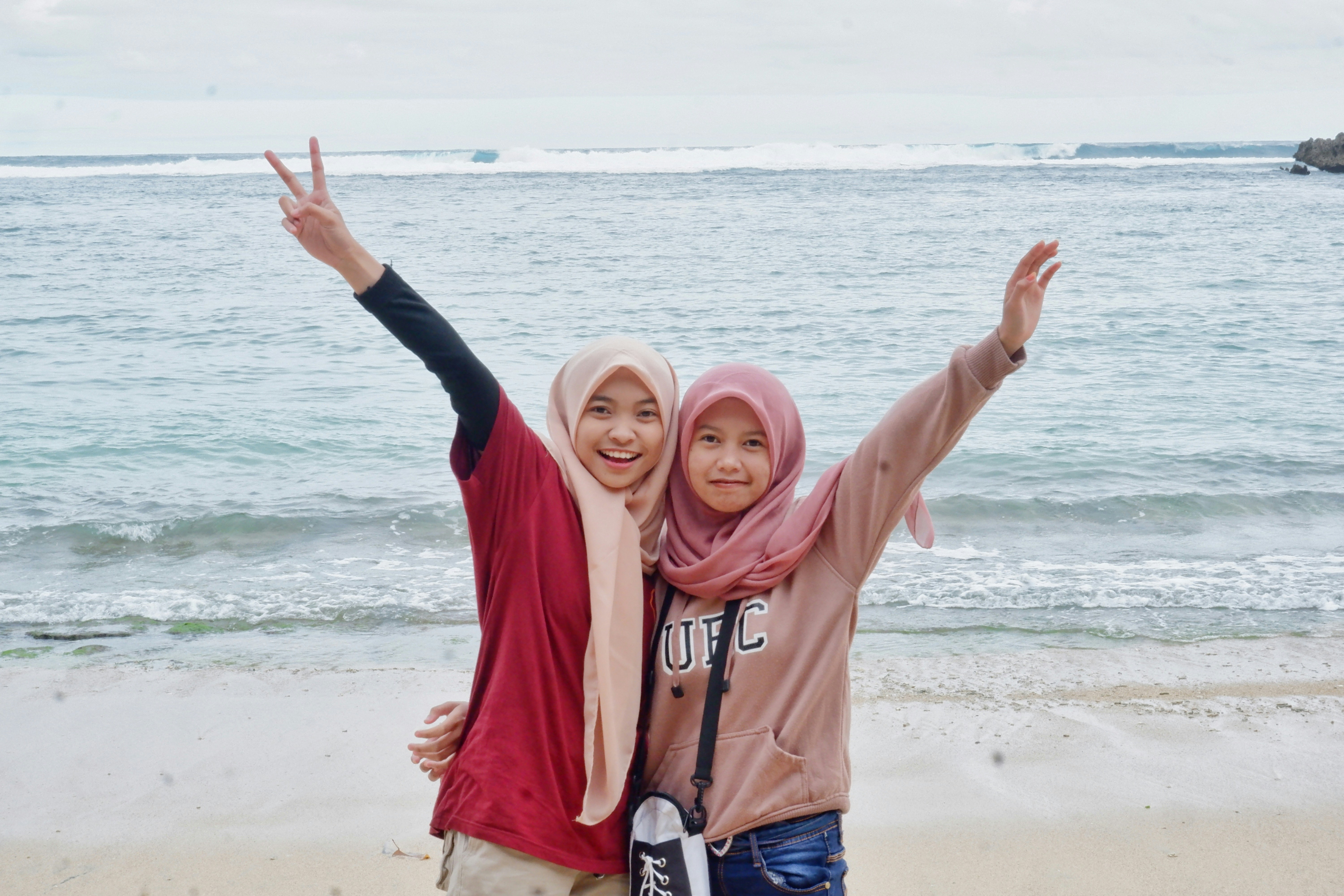 Dua wanita berjilbab tersenyum dan berpose di pantai.