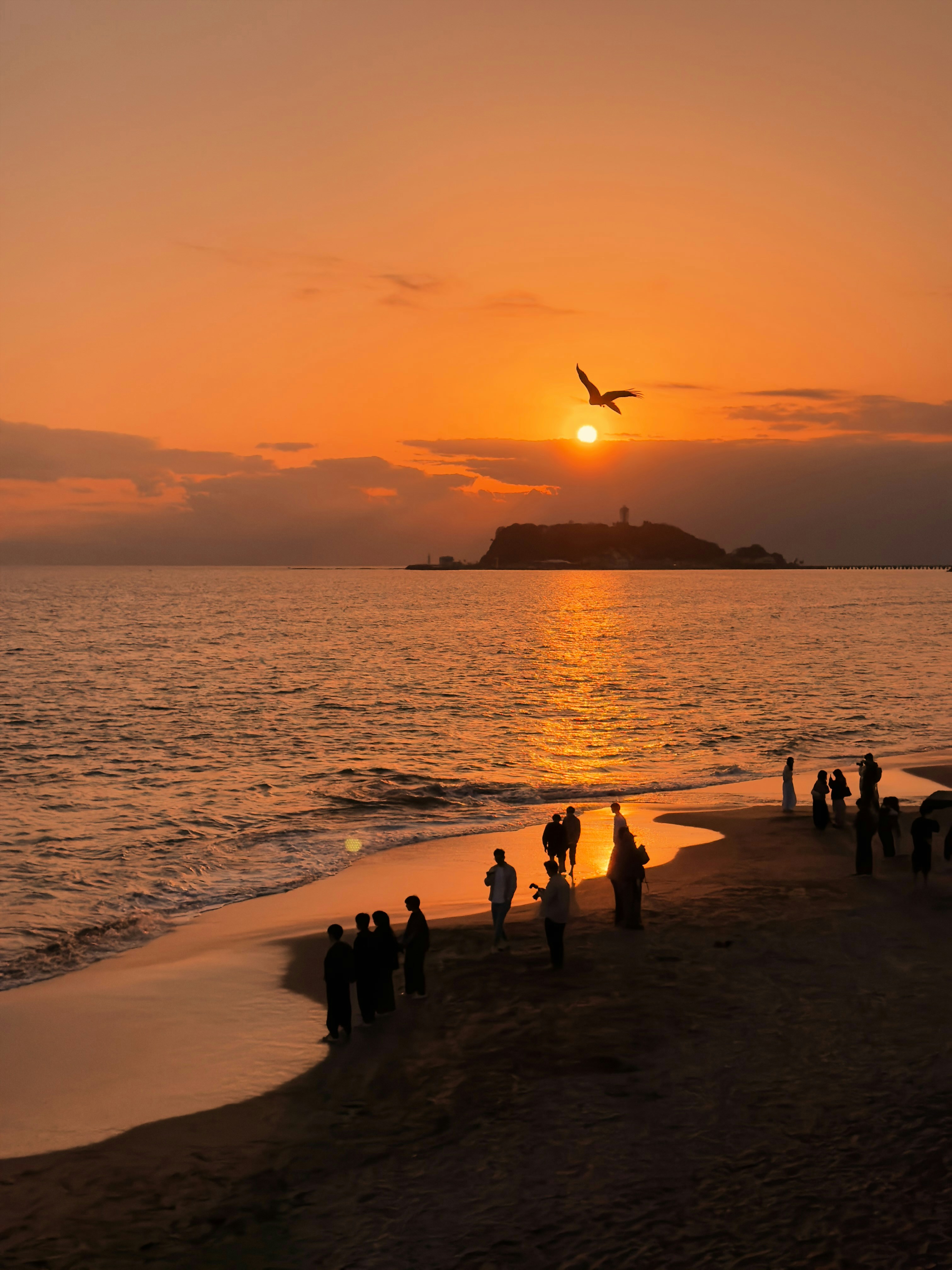 People on beach watch vibrant sunset over ocean and island.
