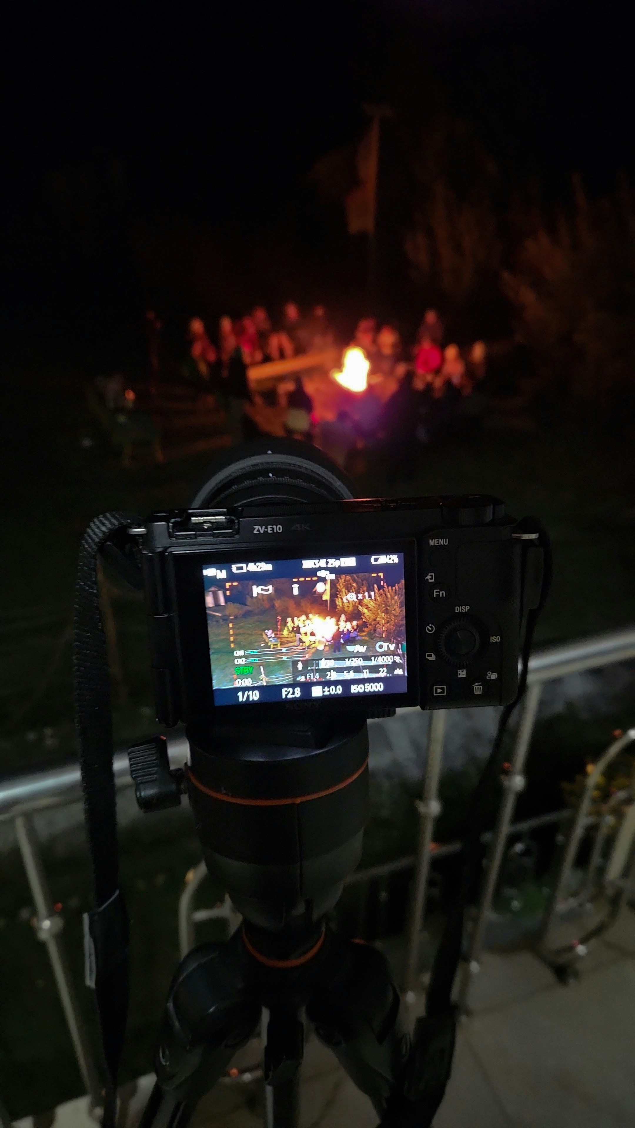 Camera on tripod filming people around a campfire at night.