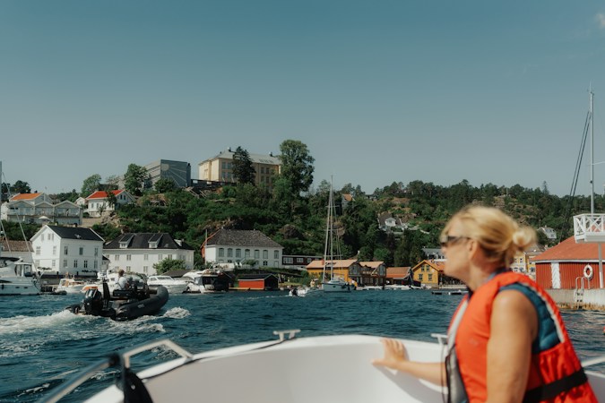 Woman in an orange foam life jacket riding in the bow of a small open boat leaving a harbor lined with white houses and moored boats