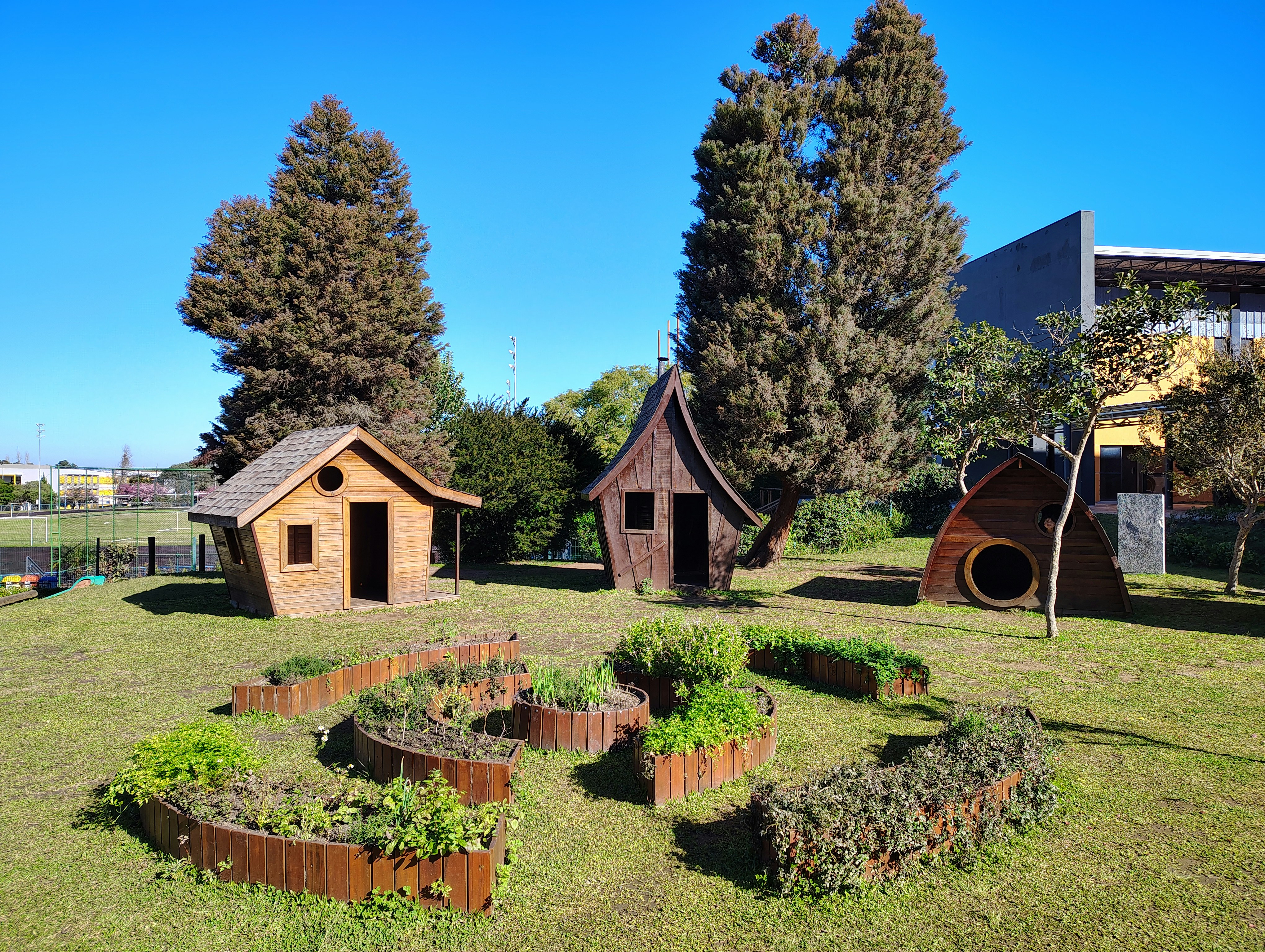 Three wooden playhouses and garden beds in a grassy park.