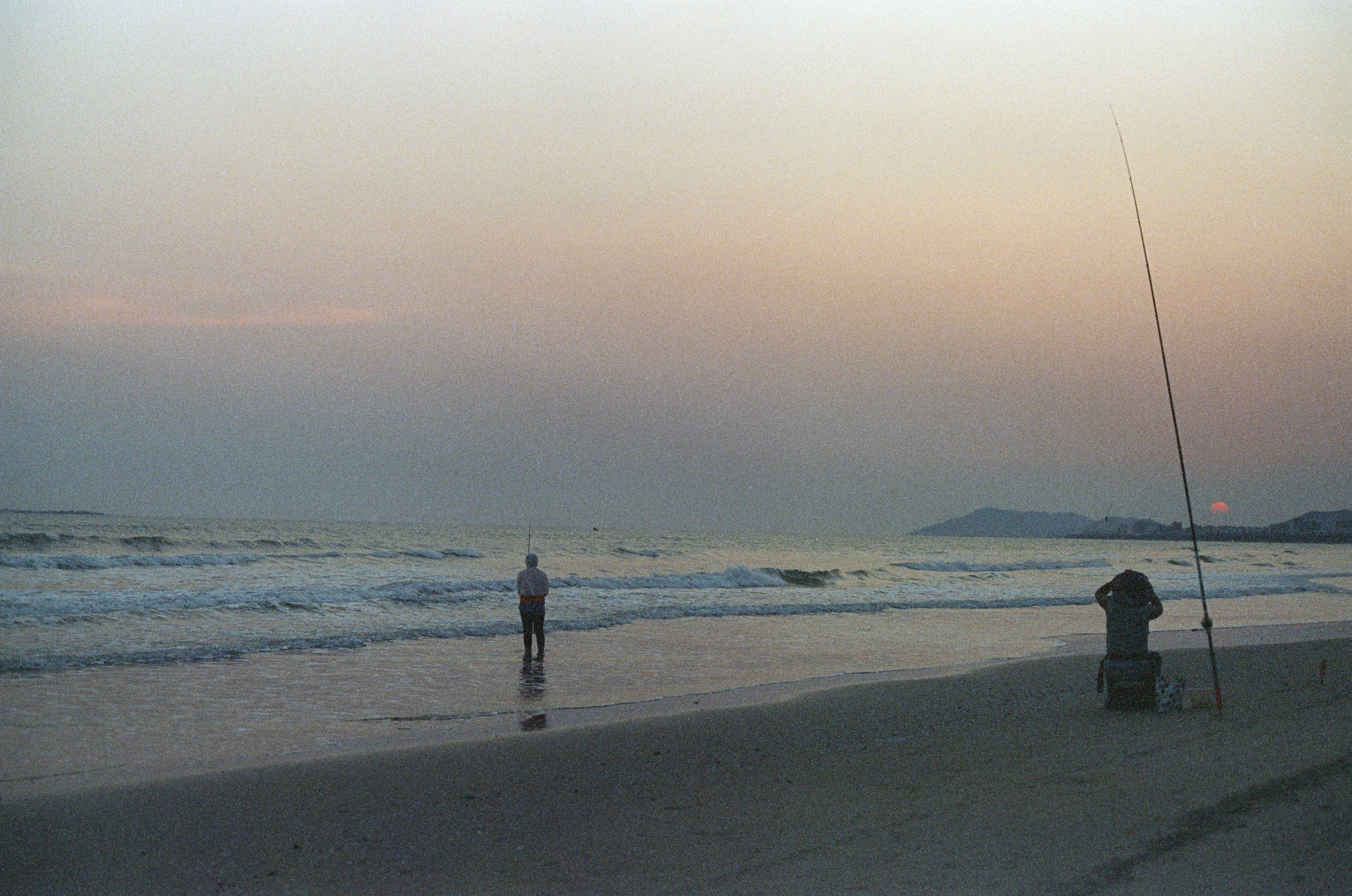 Two people fishing on a beach at sunset.