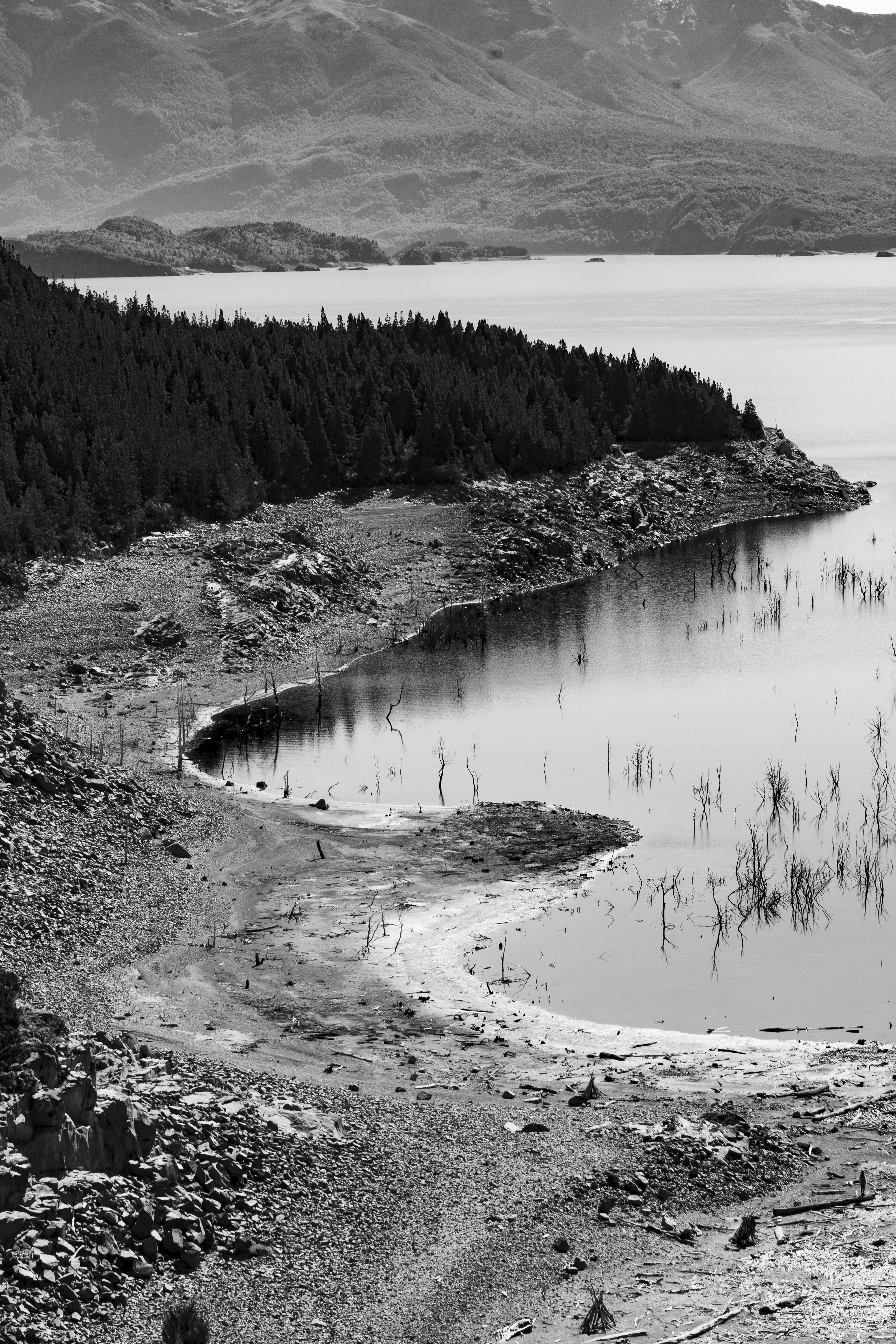 Lago em escala cinza com margem exposta e montanhas distantes.