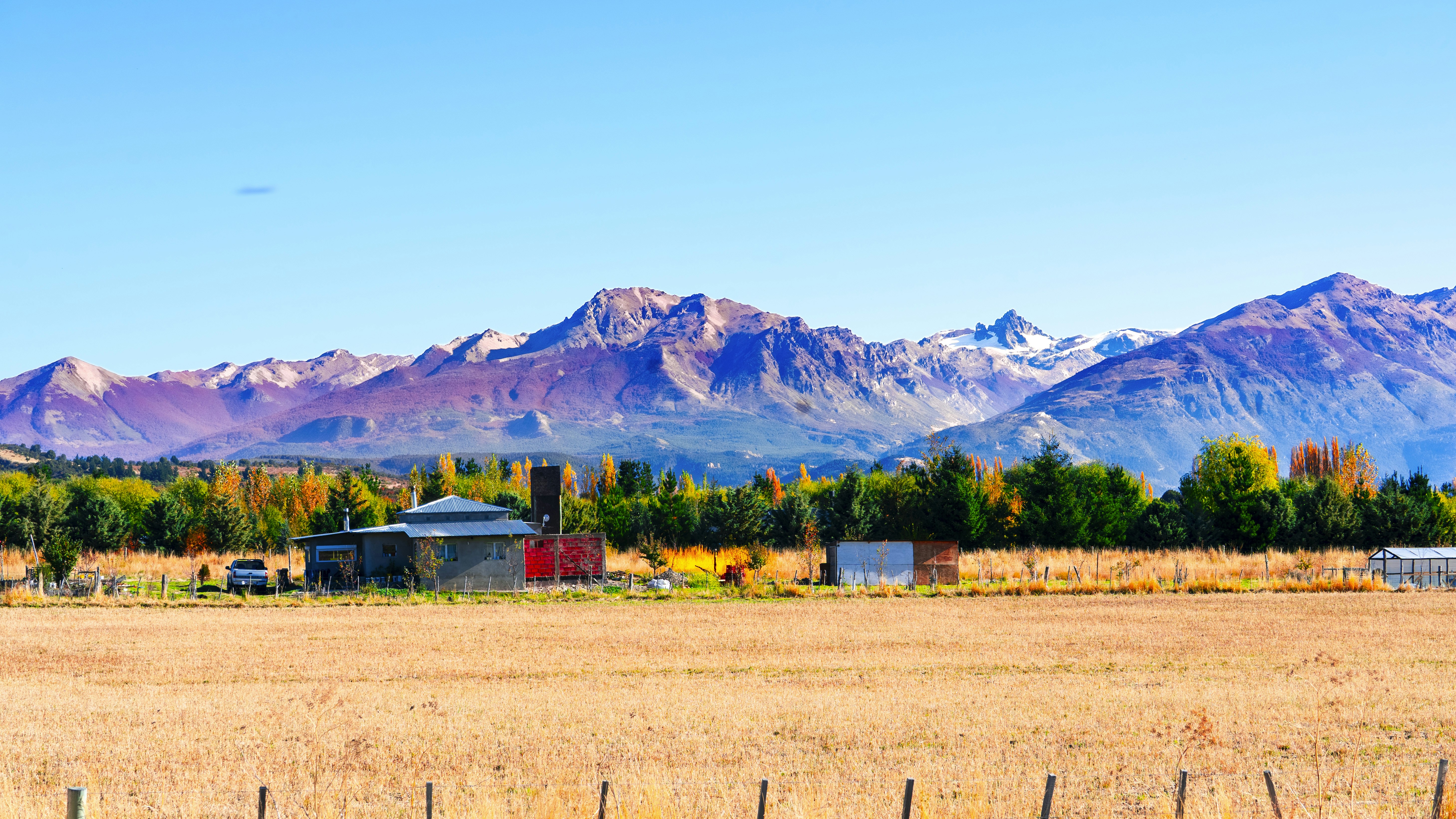 Golden field, colorful autumn trees, buildings, and majestic mountains.