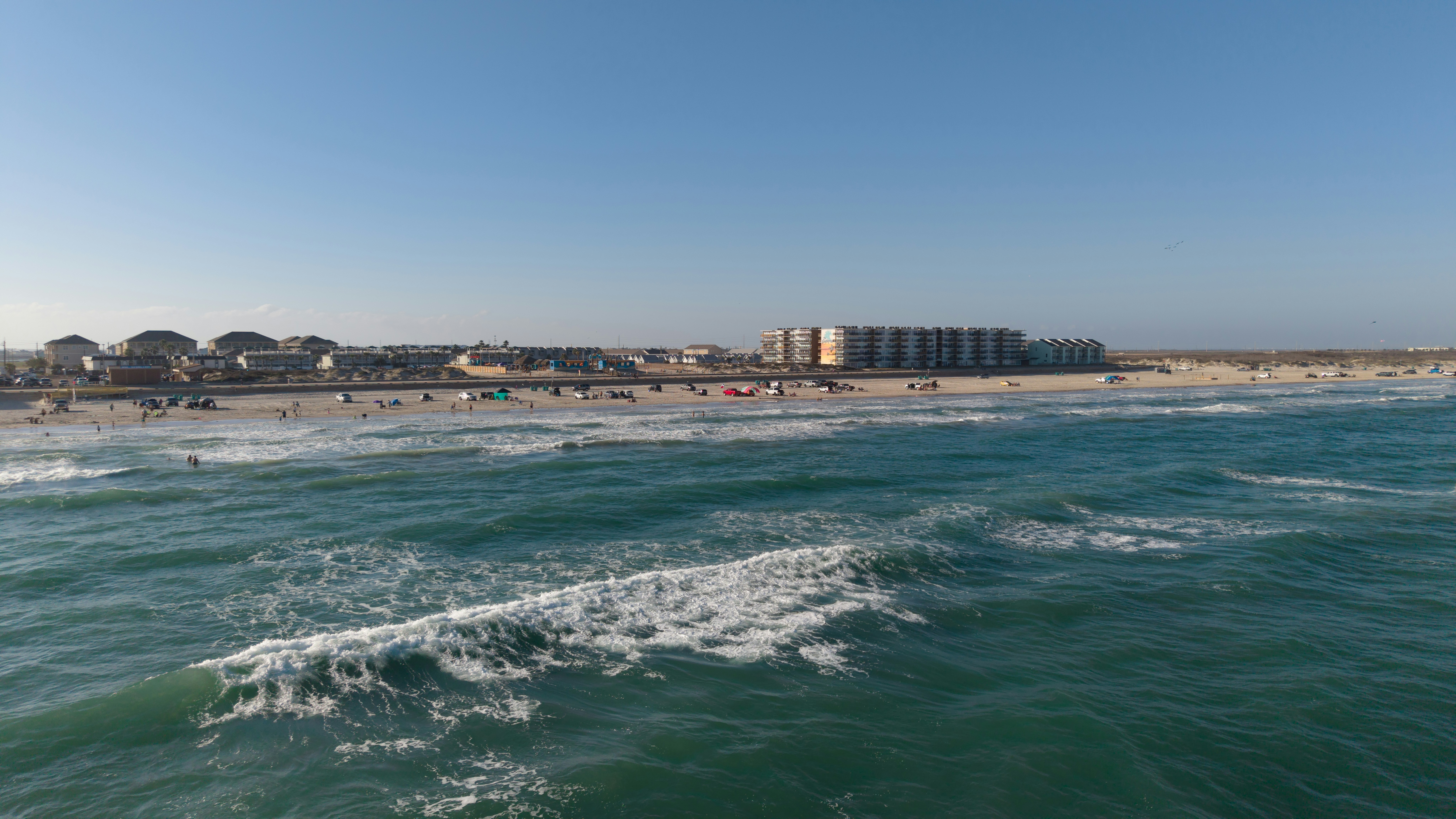 Ocean waves crash on a sandy beach with coastal buildings.