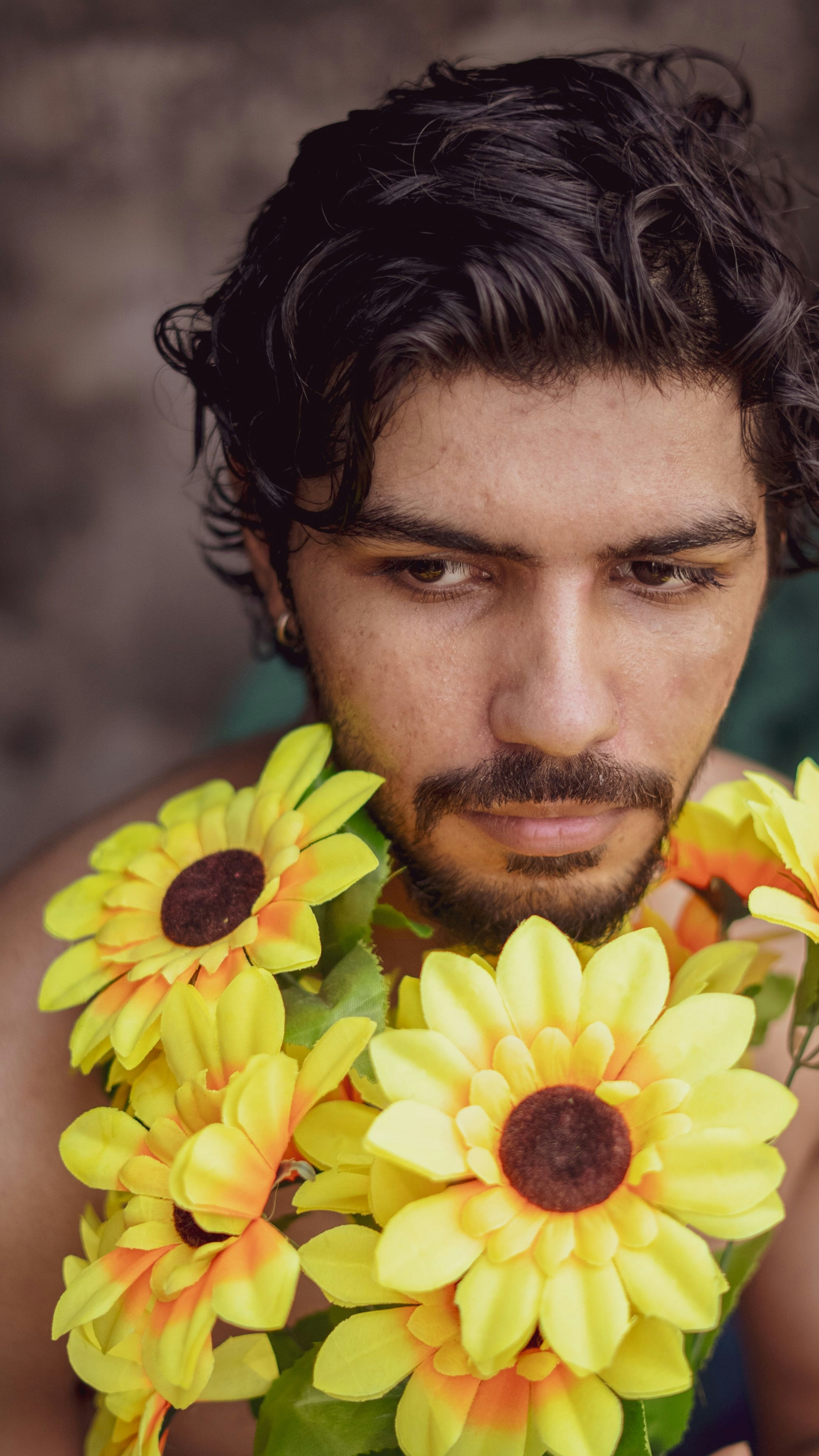 Junger Mann mit dunklem Haar und Bart, umgeben von Sonnenblumen.