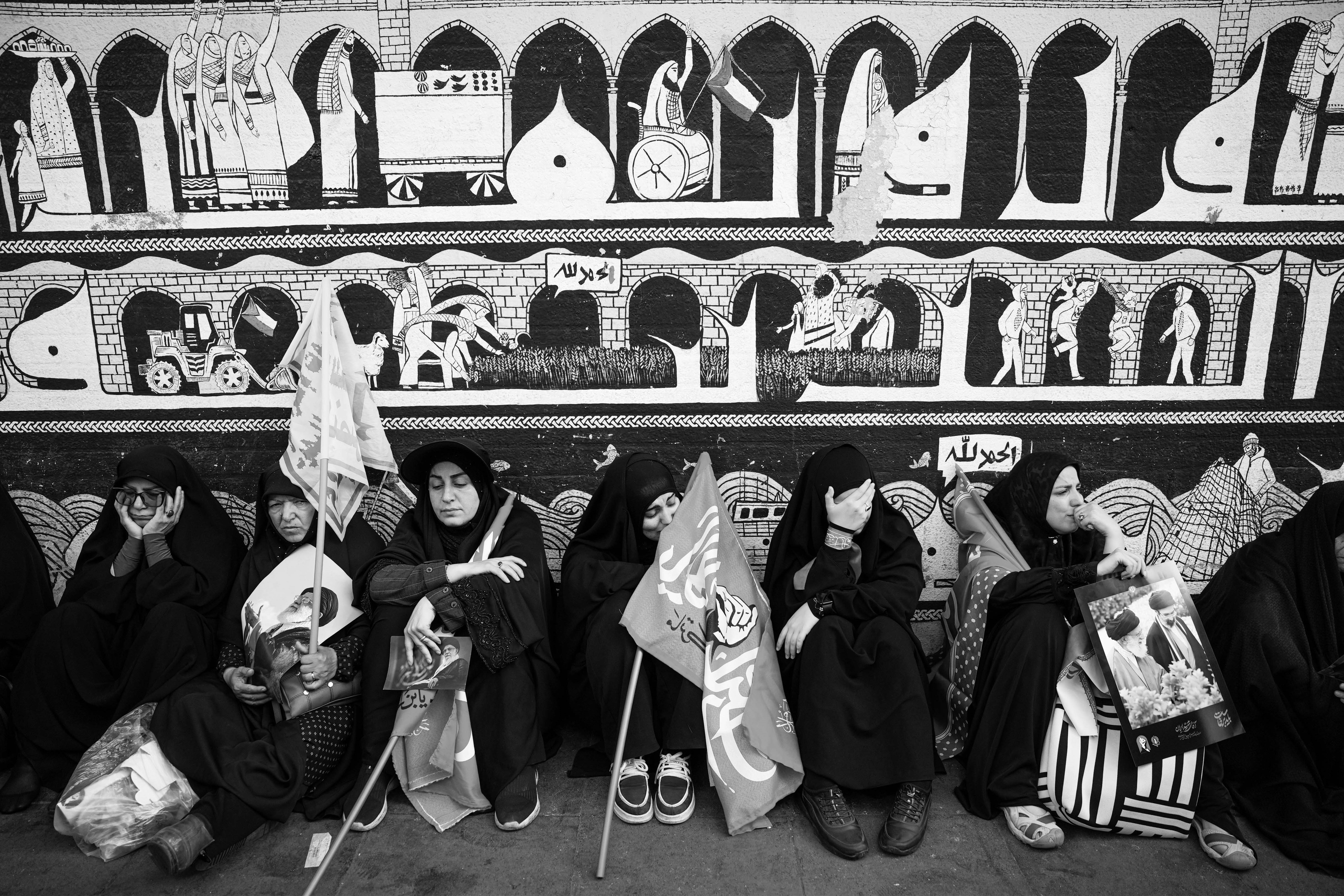 Women in hijabs sit by a decorated wall with flags