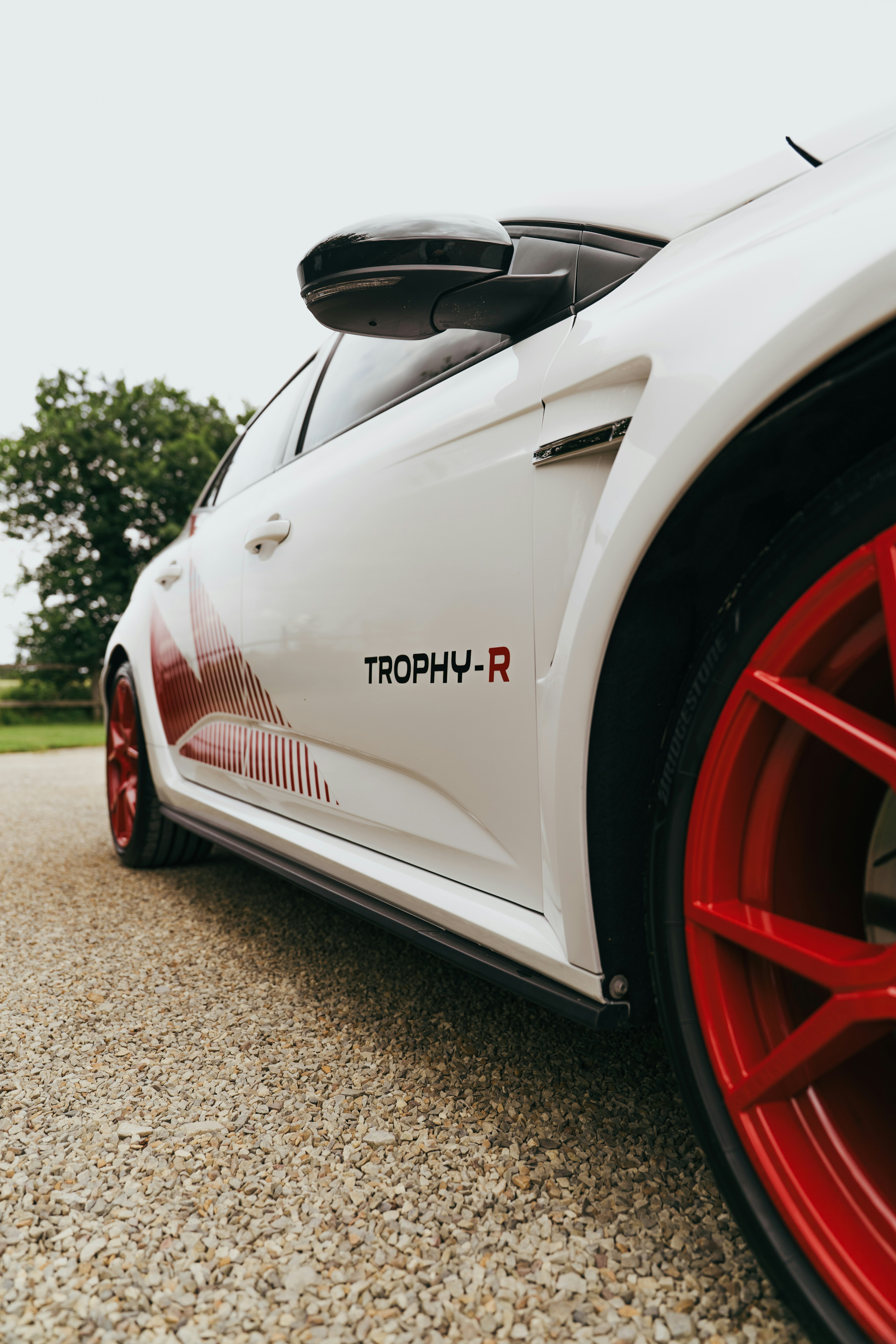 White sports car with red wheels and "trophy-r" on gravel.