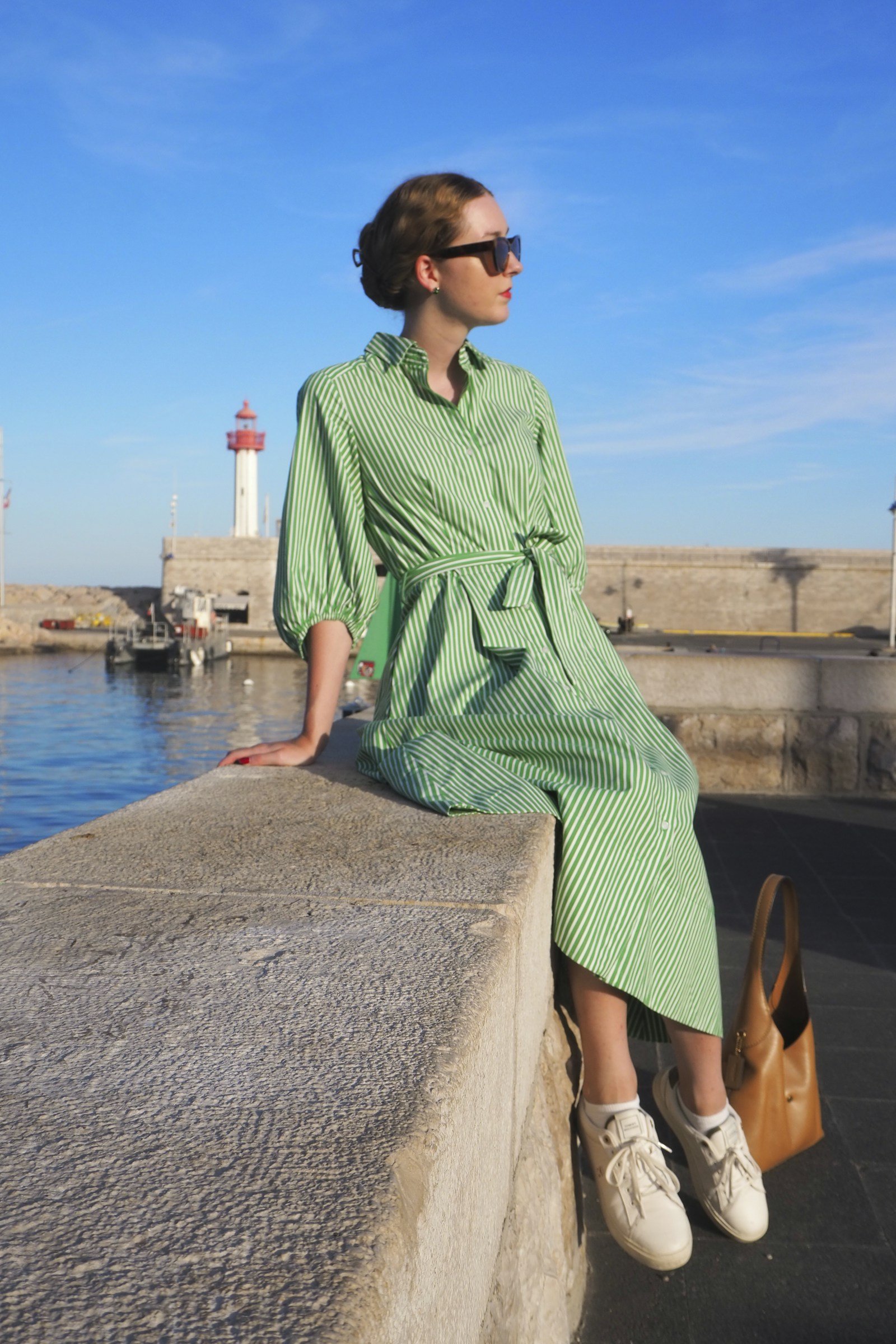 Mulher de vestido verde-sálvia sentada no cais com farol ao fundo