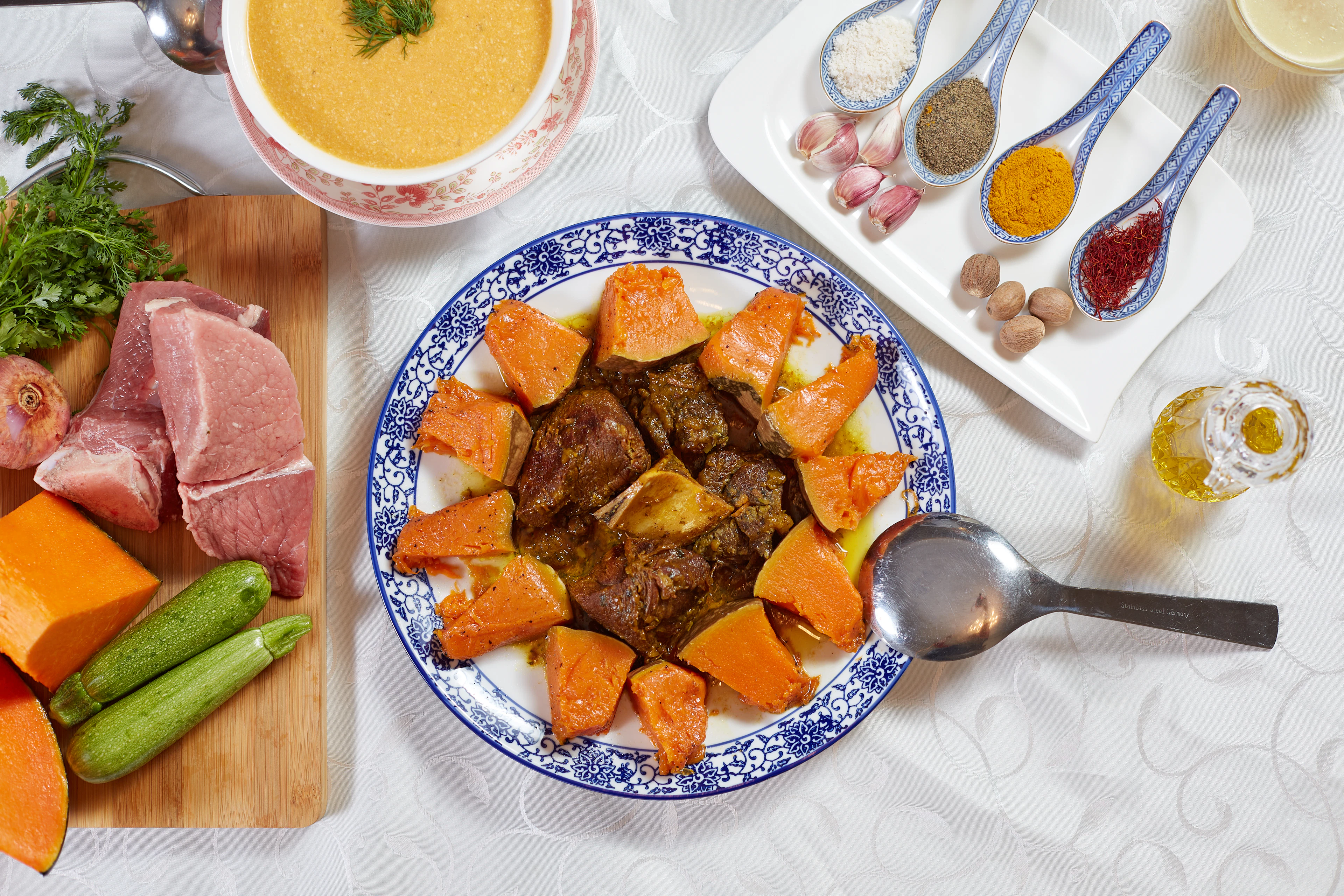 A Moroccan tagine of slow-cooked meat and pumpkin on a blue-and-white platter, photographed in Morocco beside a bowl of harira soup and spoons of ras el hanout and other spices