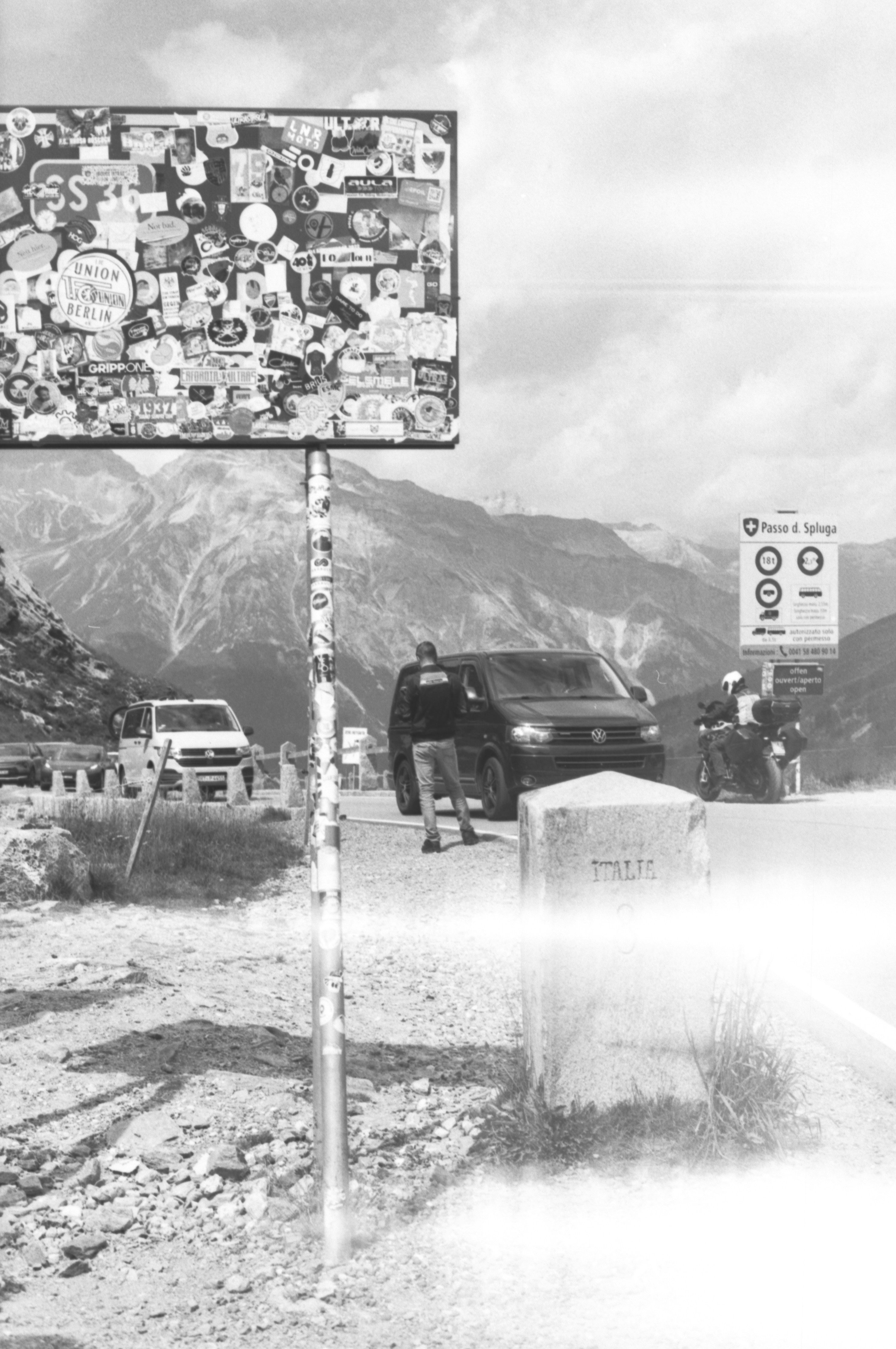 Cartello coperto di adesivi, persona e furgoni su una strada di montagna.
