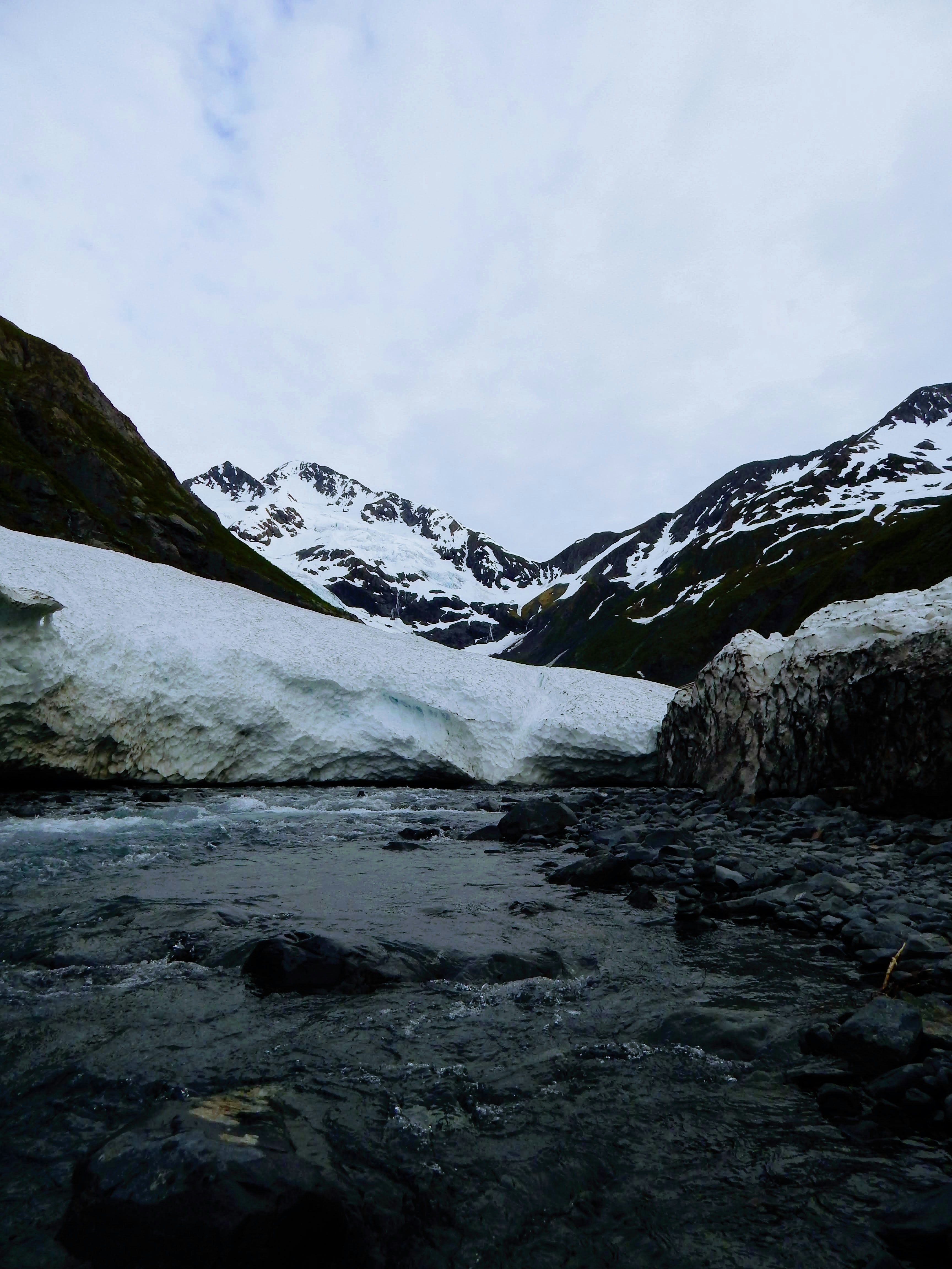 A river flows past a large glacier and snow-capped mountains.