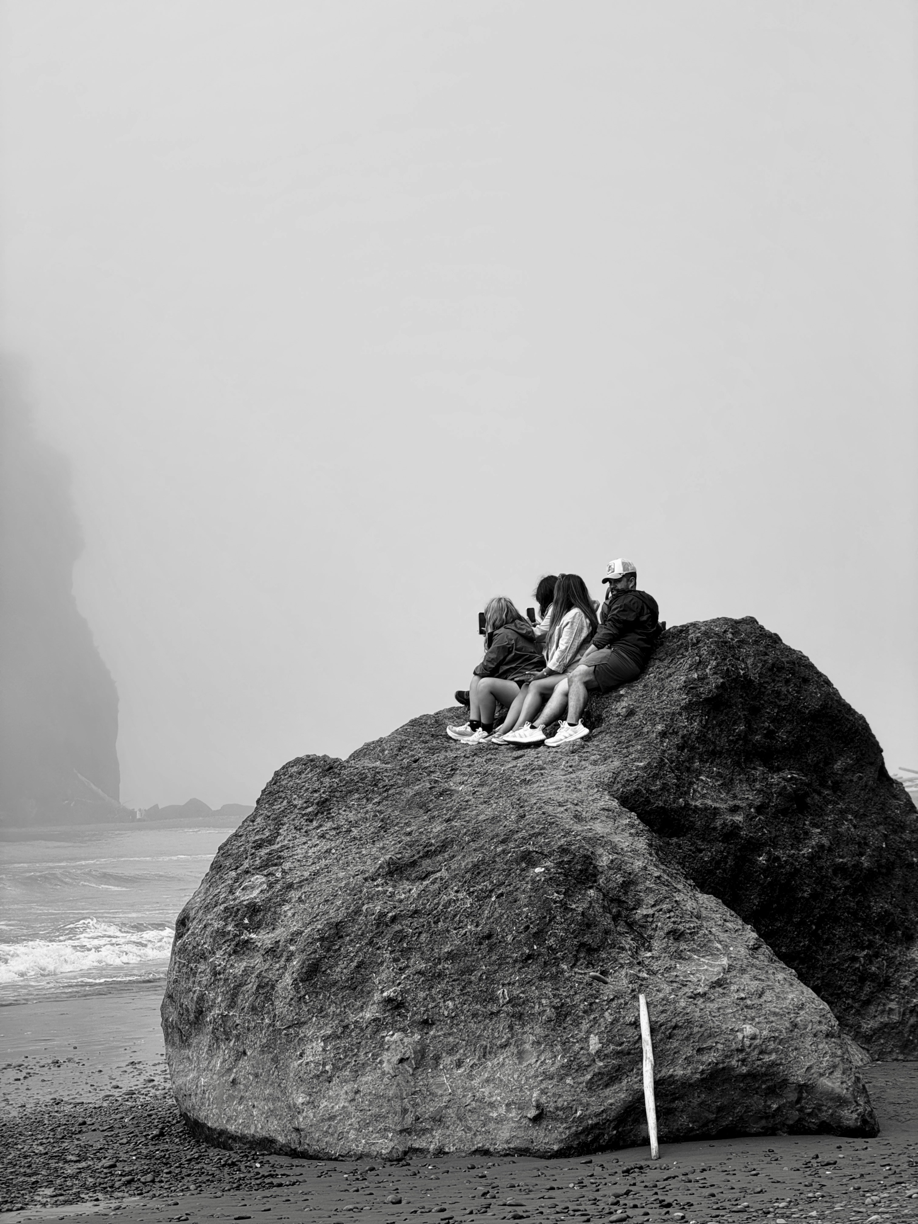 Three people sit on a large rock by the ocean.