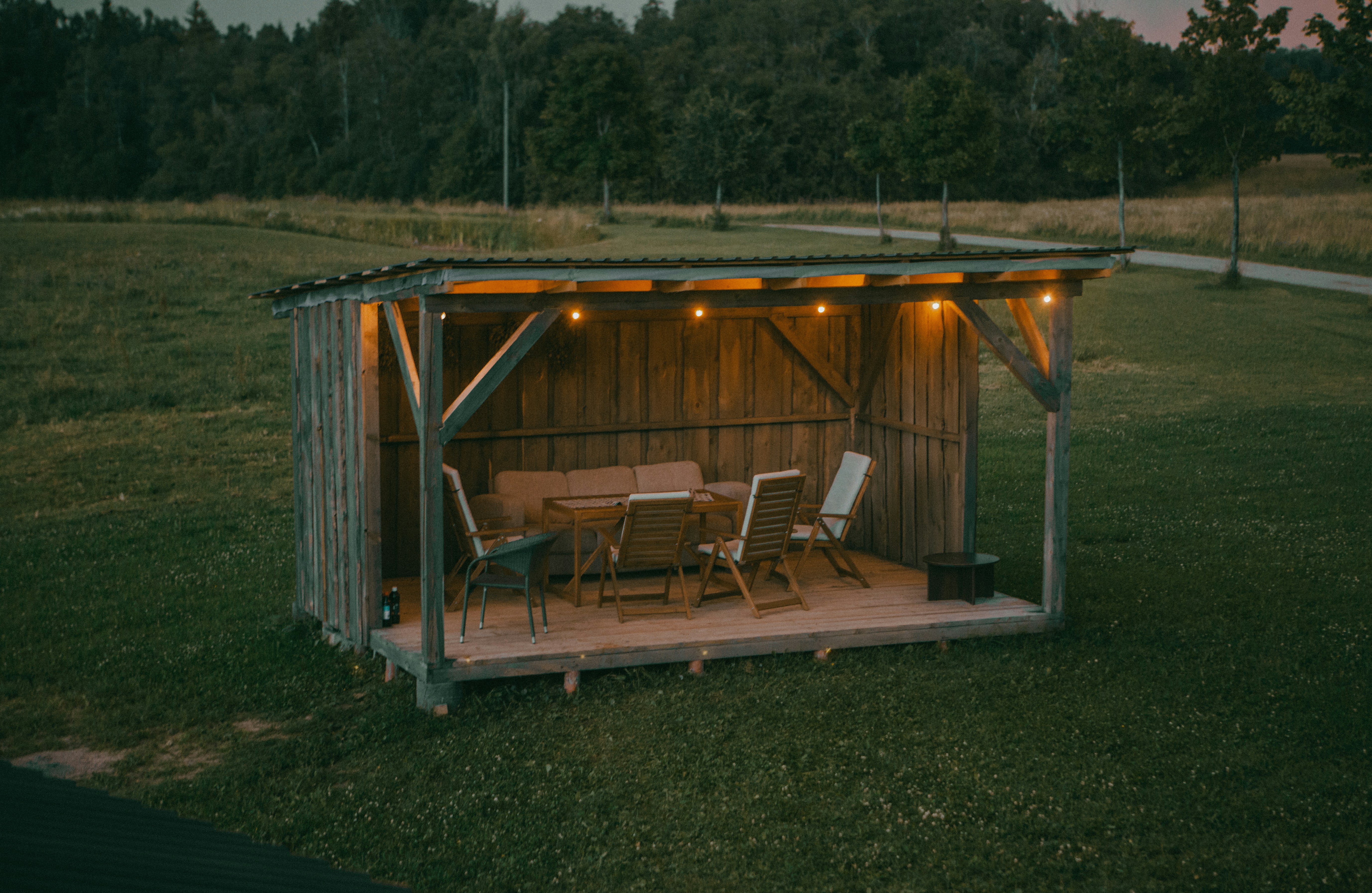 Wooden outdoor shelter with string lights and furniture at dusk.