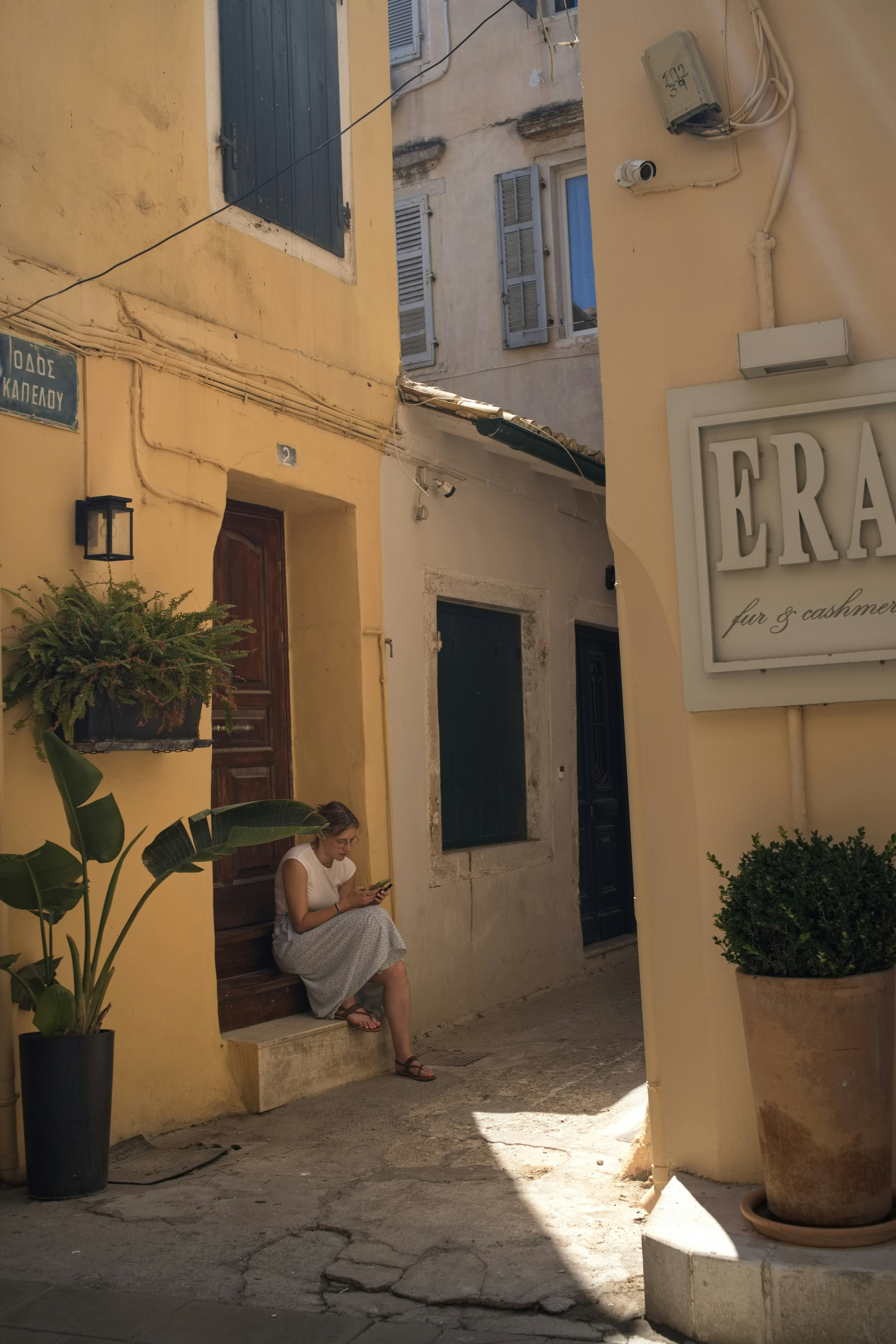 A woman sits on steps in a sunlit, narrow alleyway.