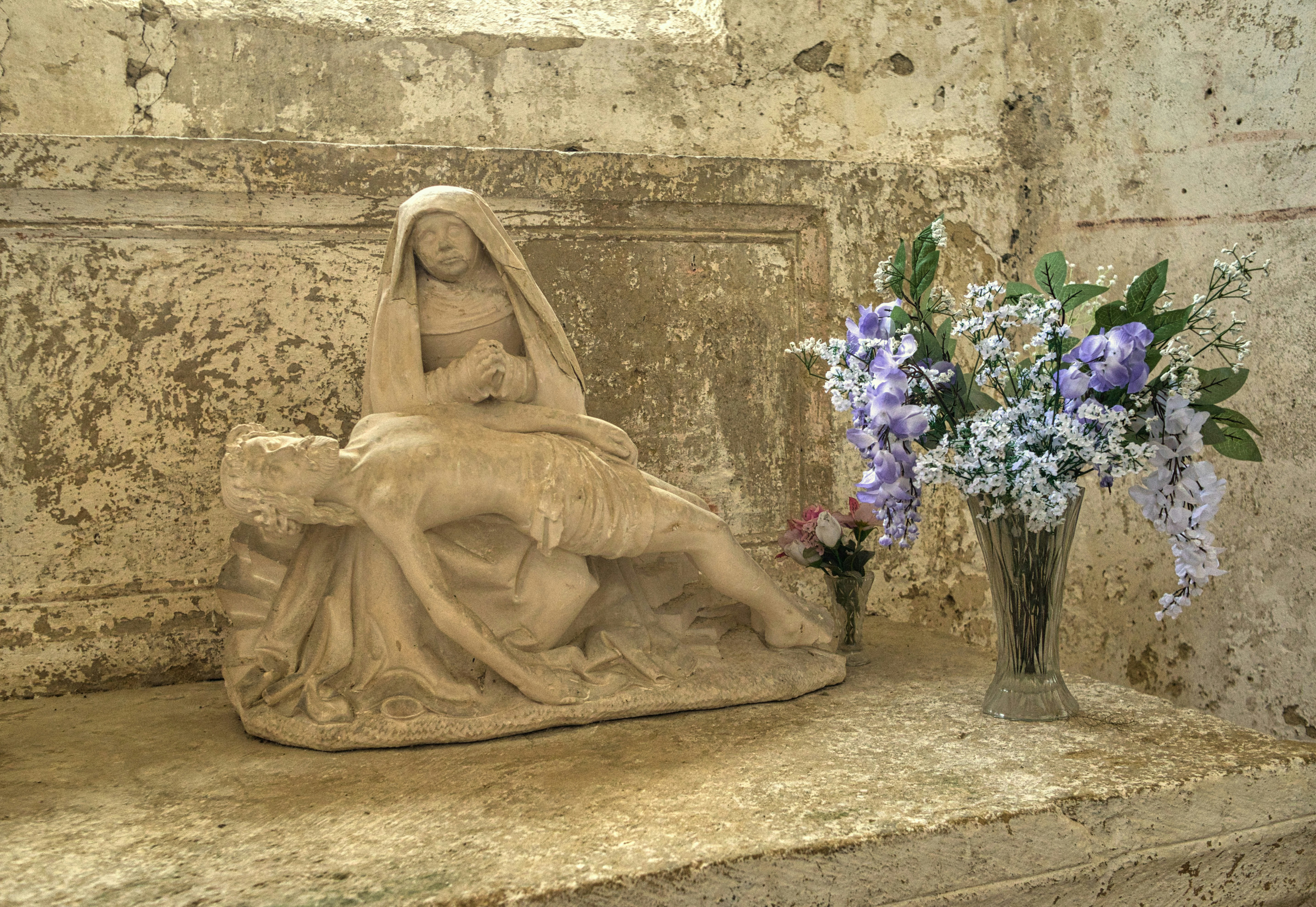Pietà statue with fresh flowers on an old stone shelf.