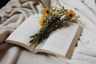 An open book with wildflowers rests on a soft blanket.