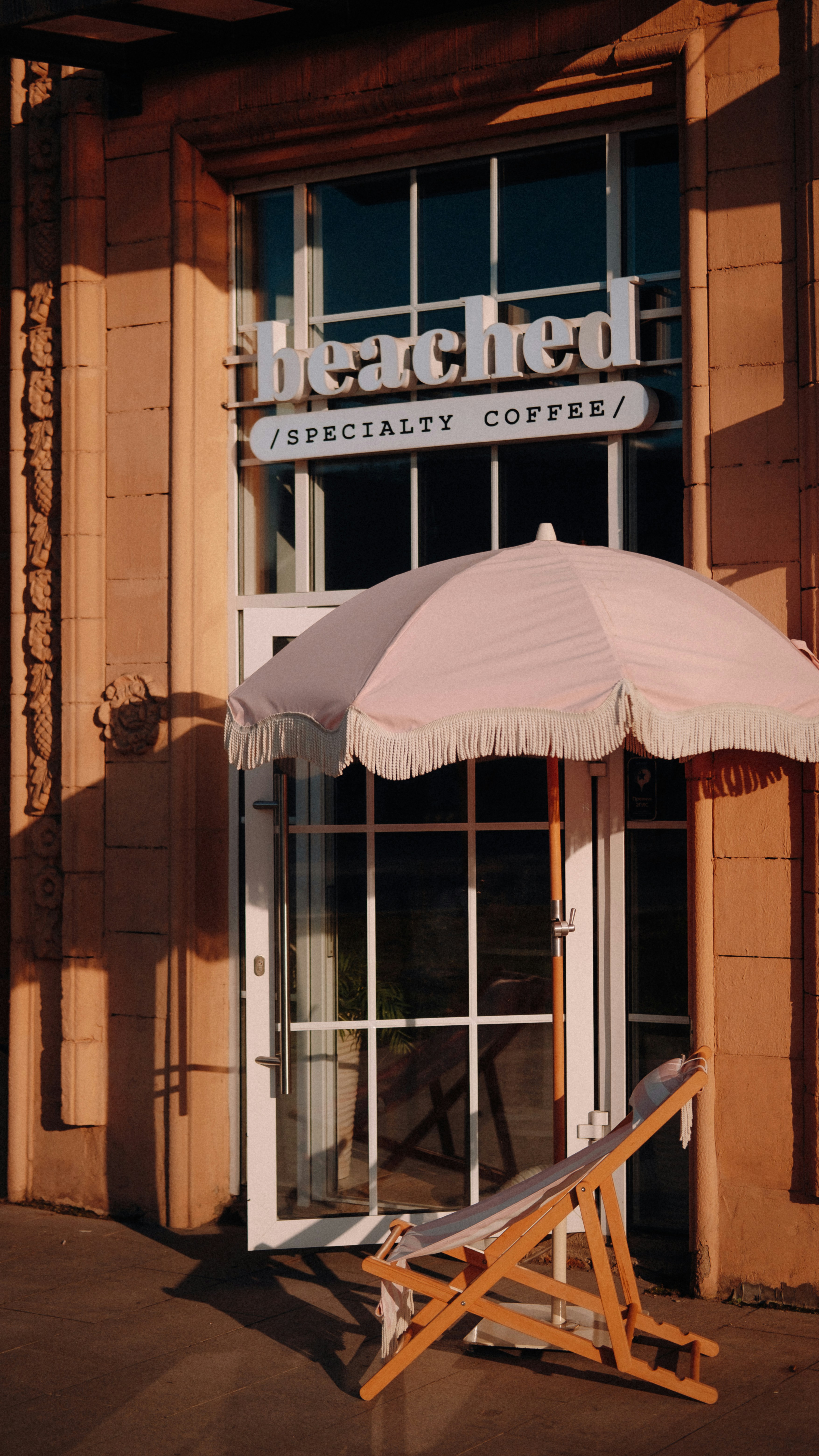 A cafe entrance with a pink umbrella and deck chair.