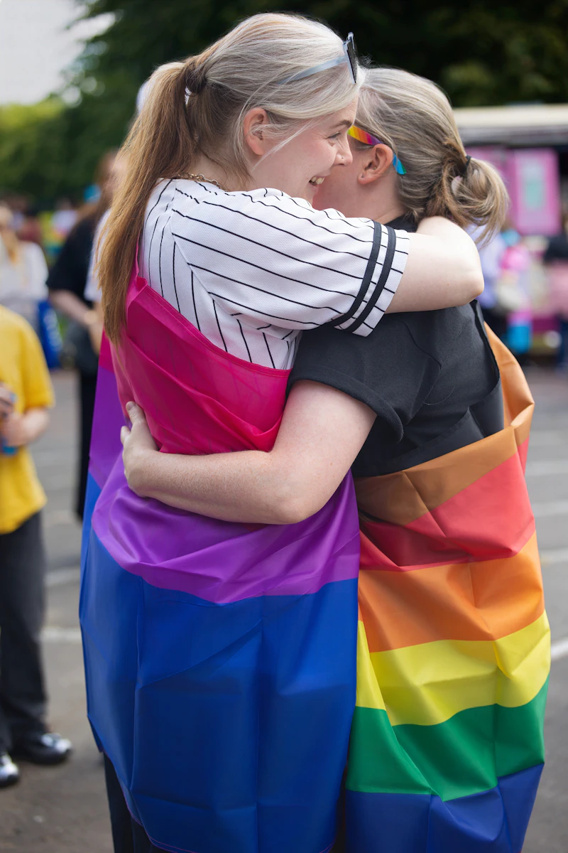 Two people wrapped together in Pride flags, hugging at an outdoor celebration