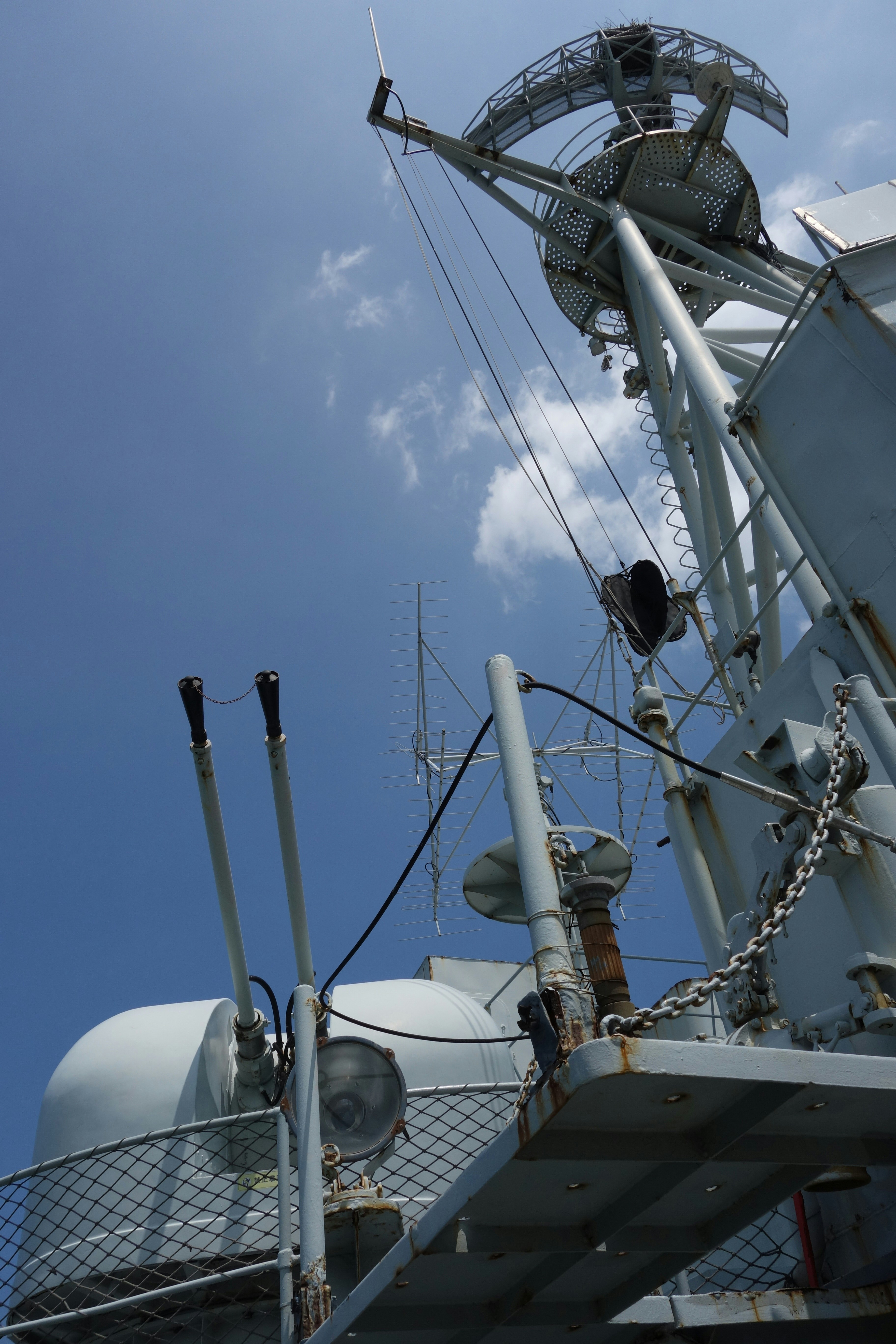 Naval ship's mast with radar and guns under blue sky.