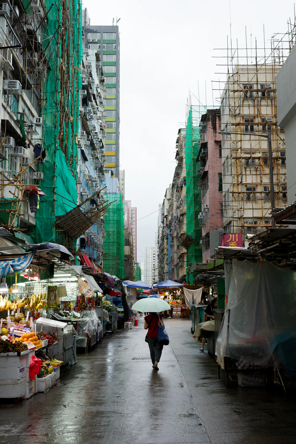 A person with a pale umbrella walks past fruit stalls on a rainy day