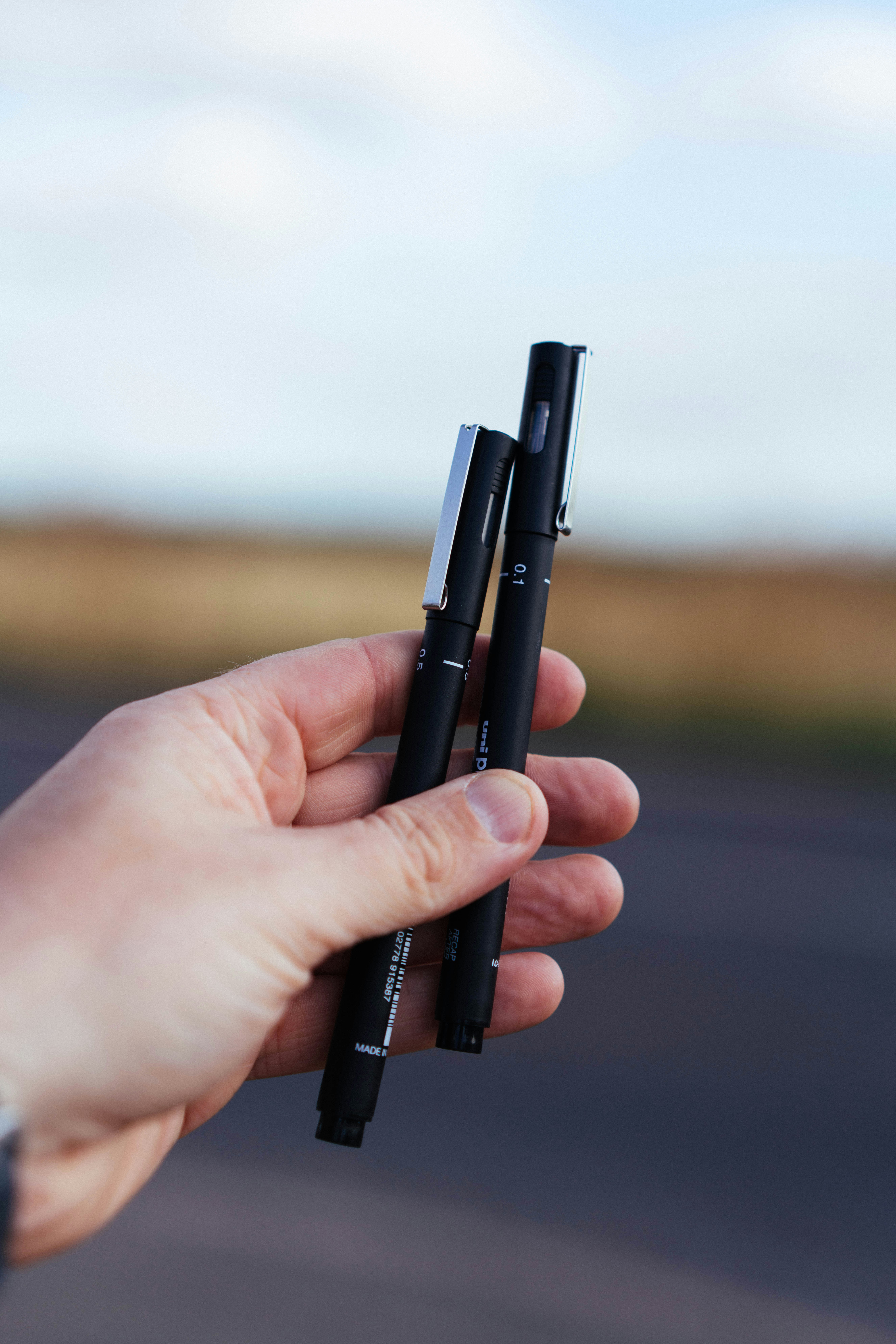 Hand holding two black pens outdoors.