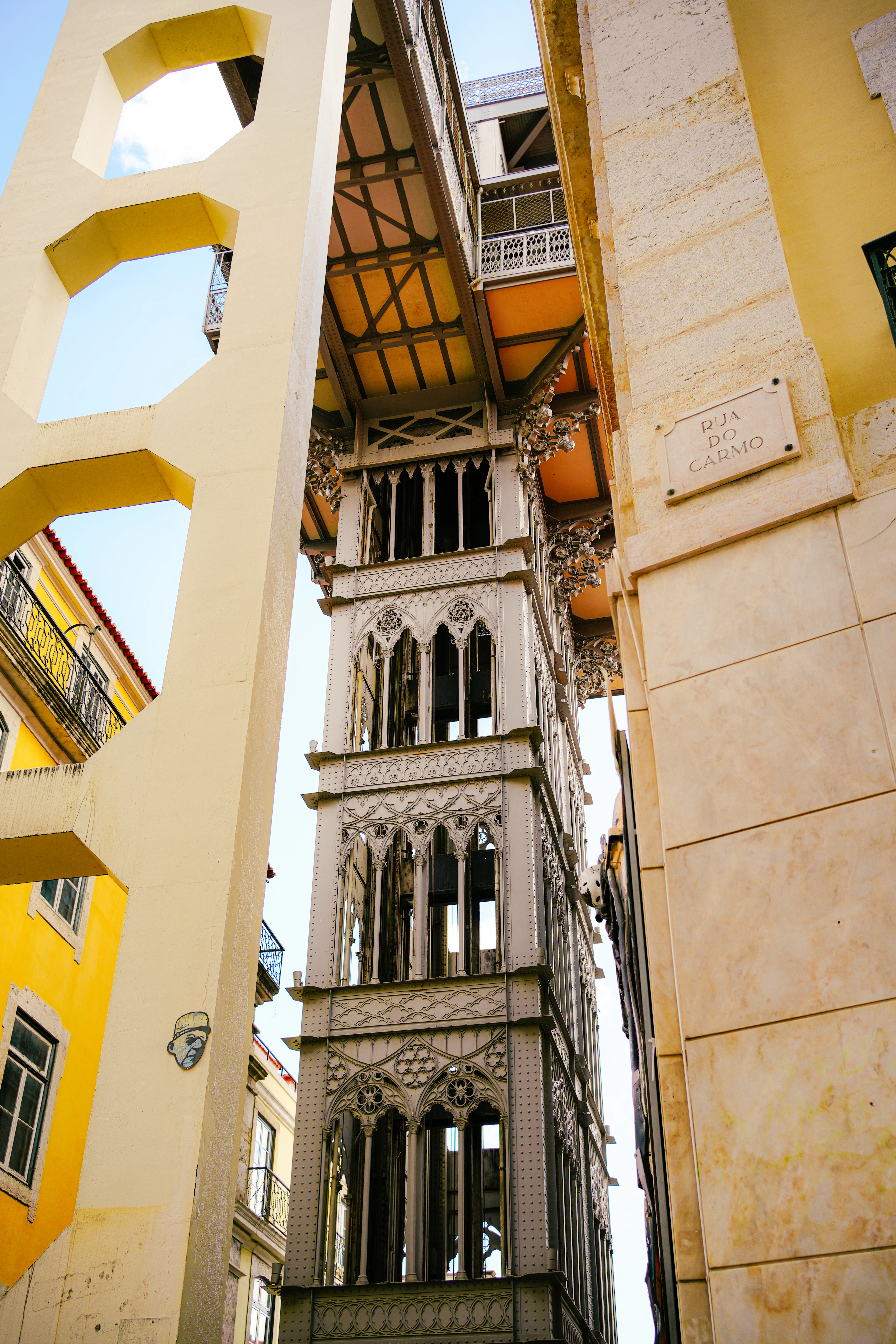 The wrought-iron Santa Justa Lift rising in the narrow gap between sunlit yellow and cream buildings on Rua do Carmo in Lisbon, its orange-painted underside and Gothic tracery seen from directly below, the carved stone street plaque on the wall beside it