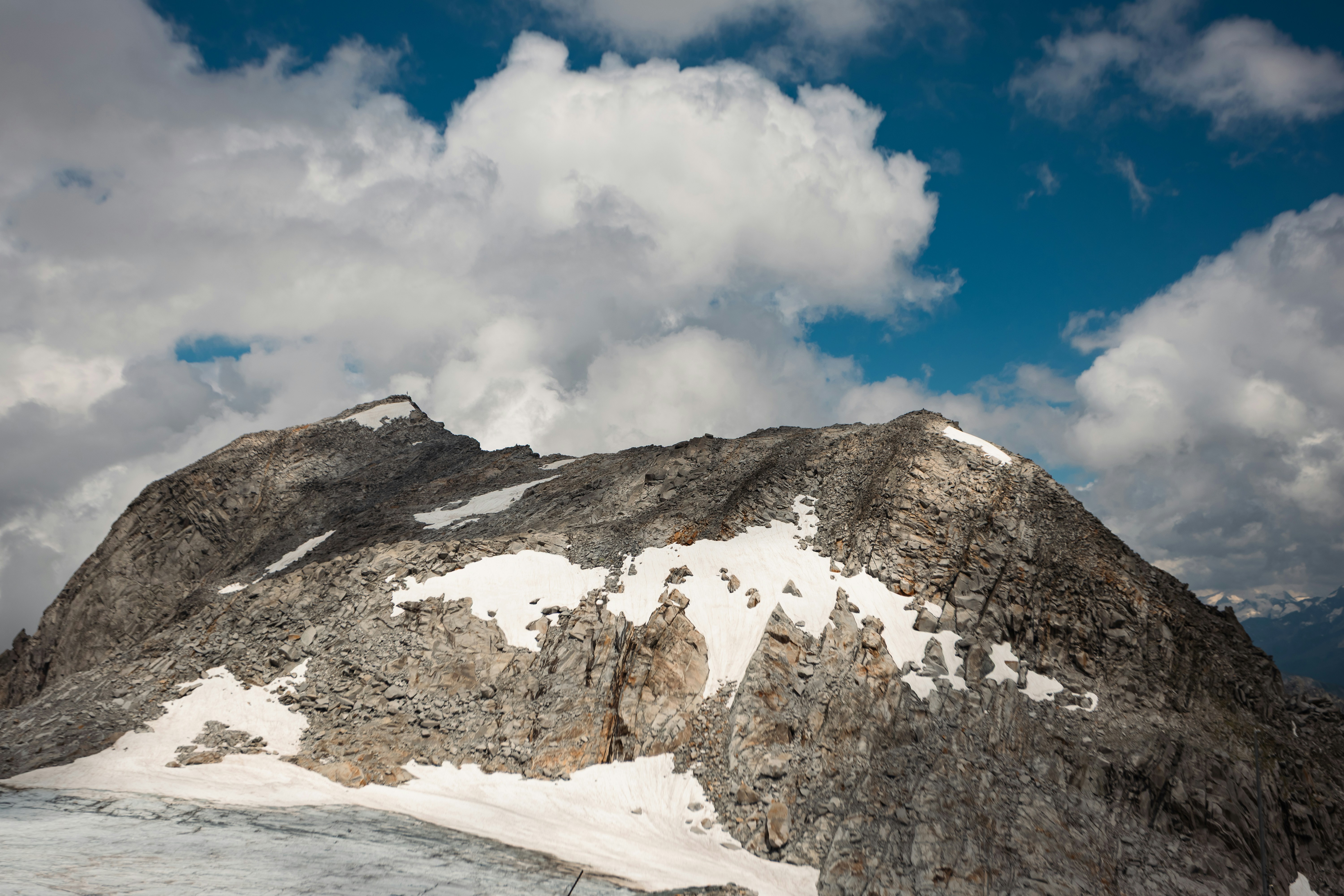 Rocky mountain peak with snow under a cloudy blue sky.