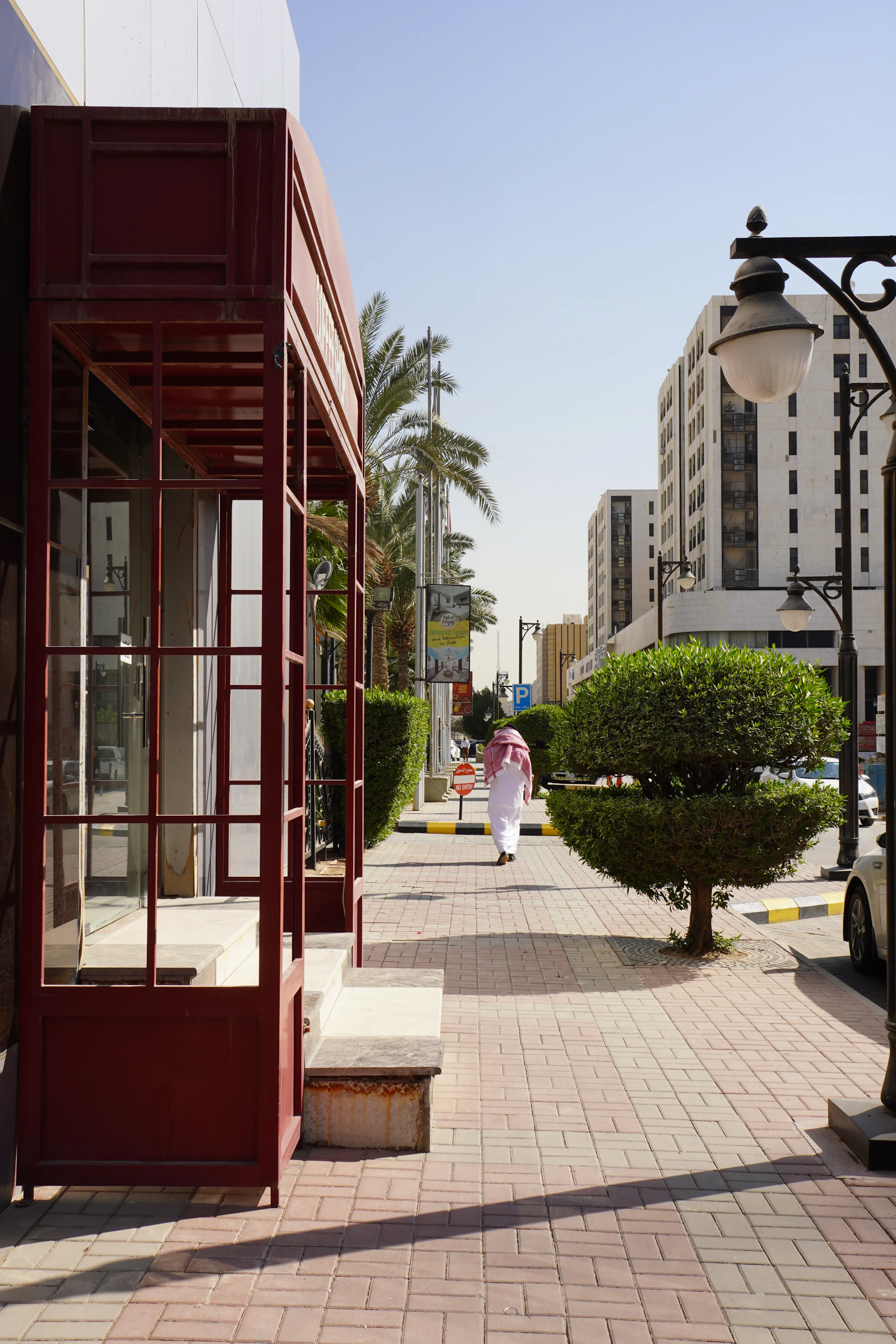 A man in a white thobe walking along a sunny, tree-lined pavement