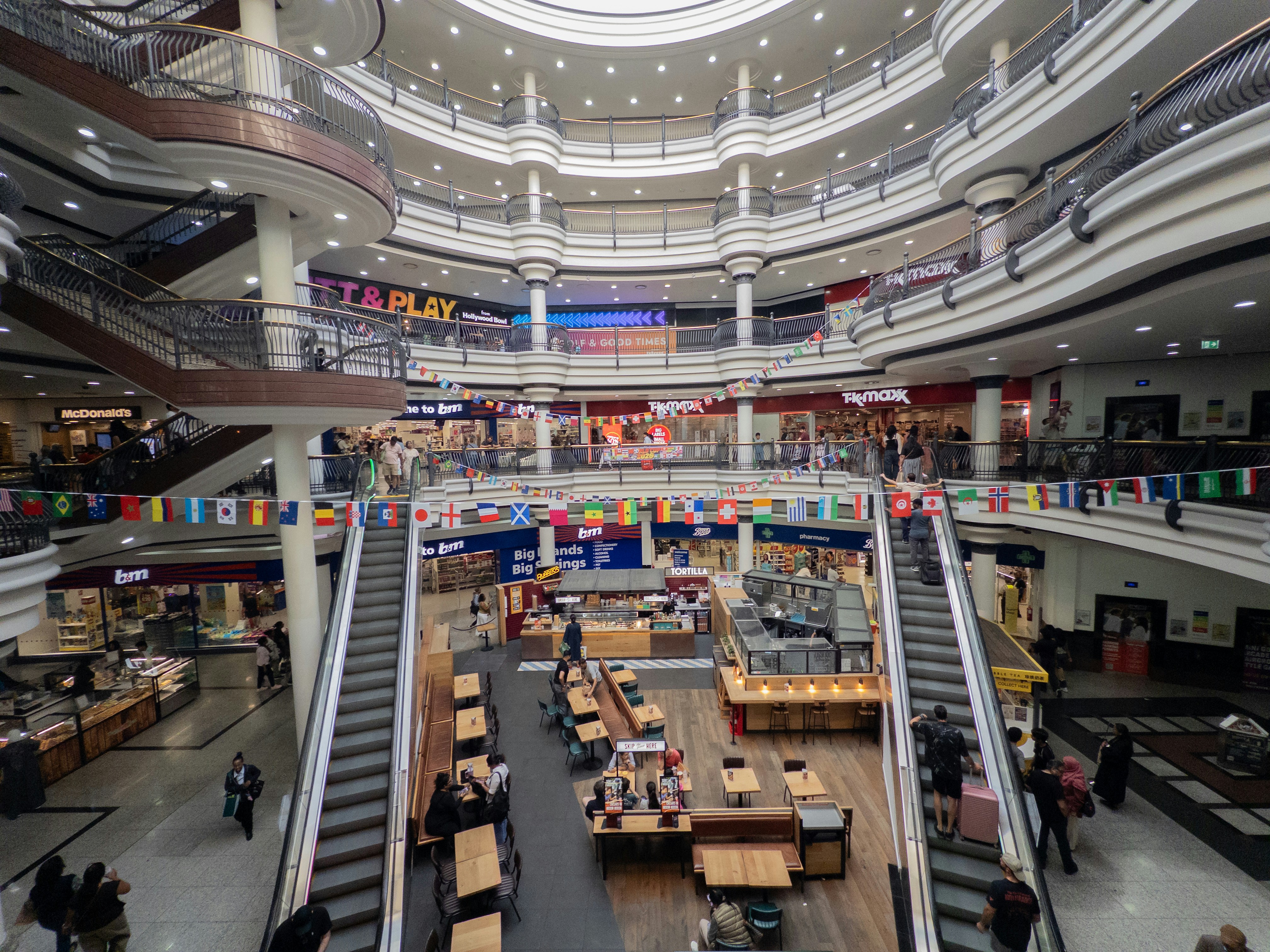 Busy multi-story shopping mall interior with escalators and domed ceiling.
