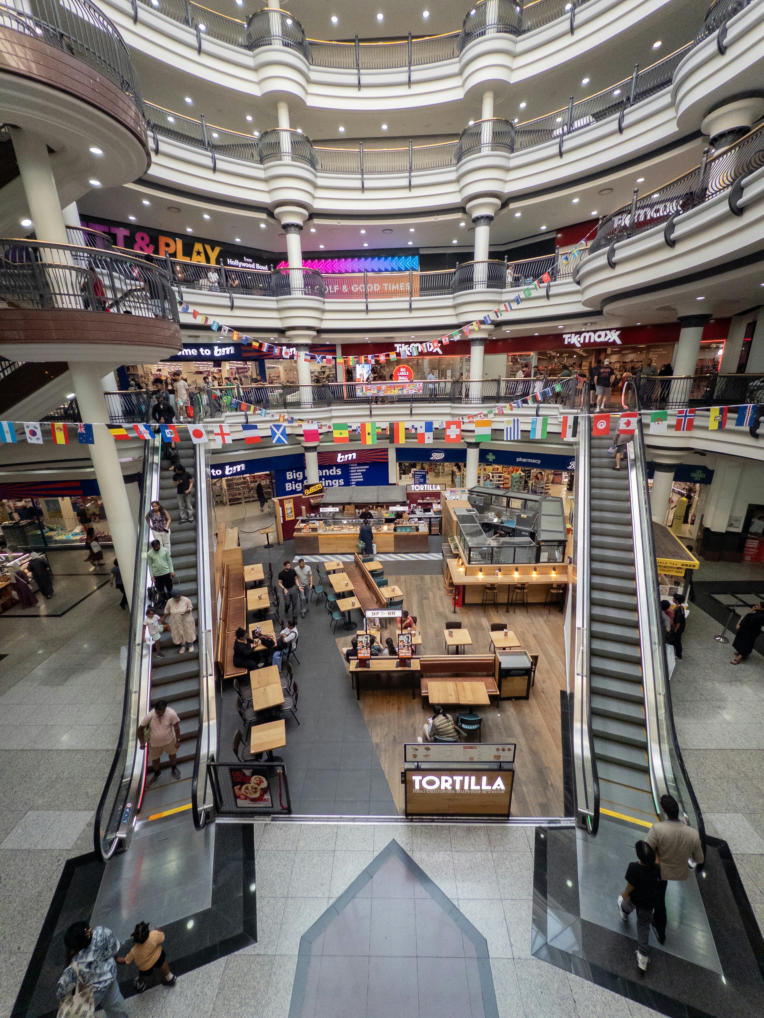 Bustling multi-level shopping mall interior with escalators and stores.