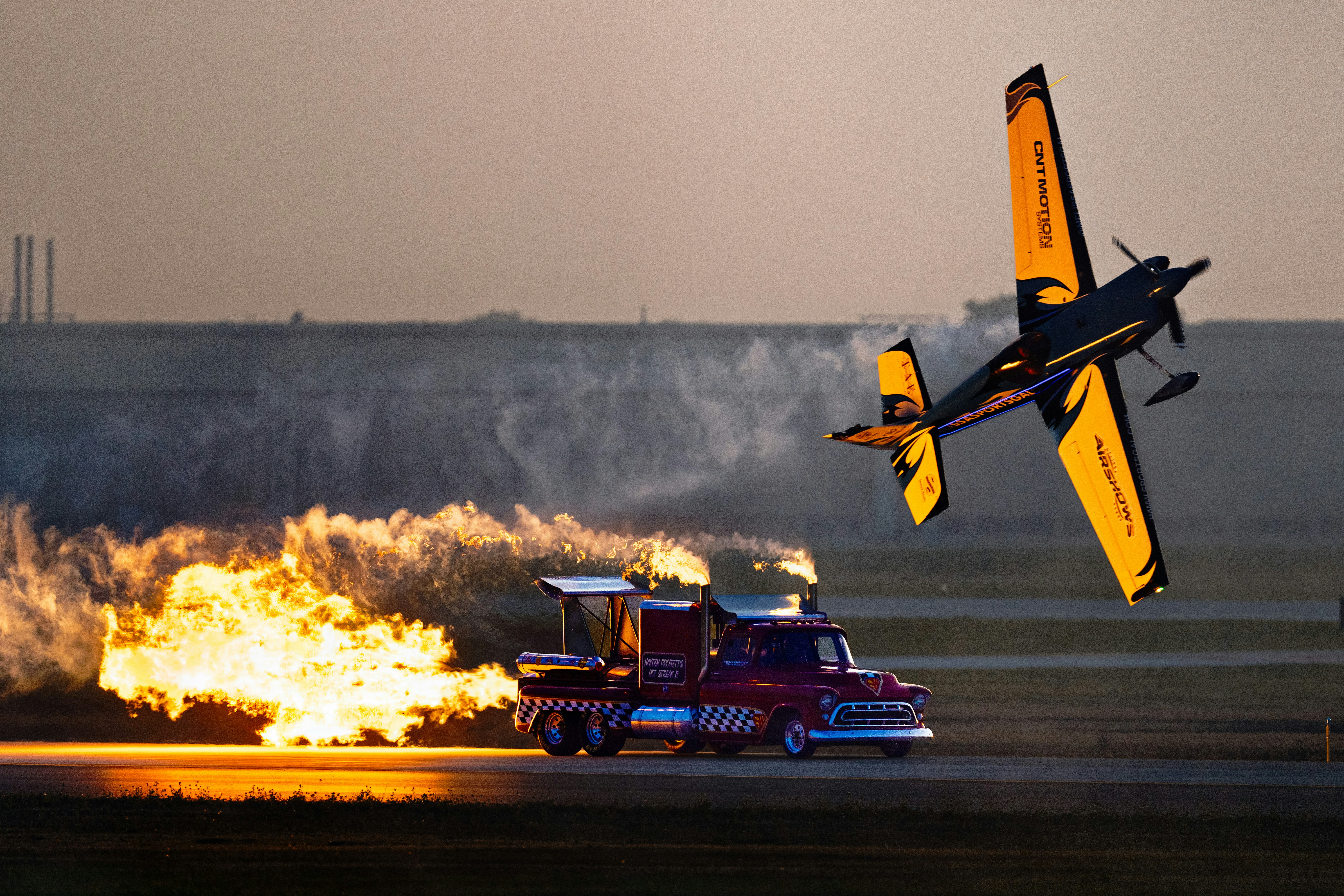 Avion cascadeur volant la tête en bas au-dessus d’un camion à réaction en flammes.