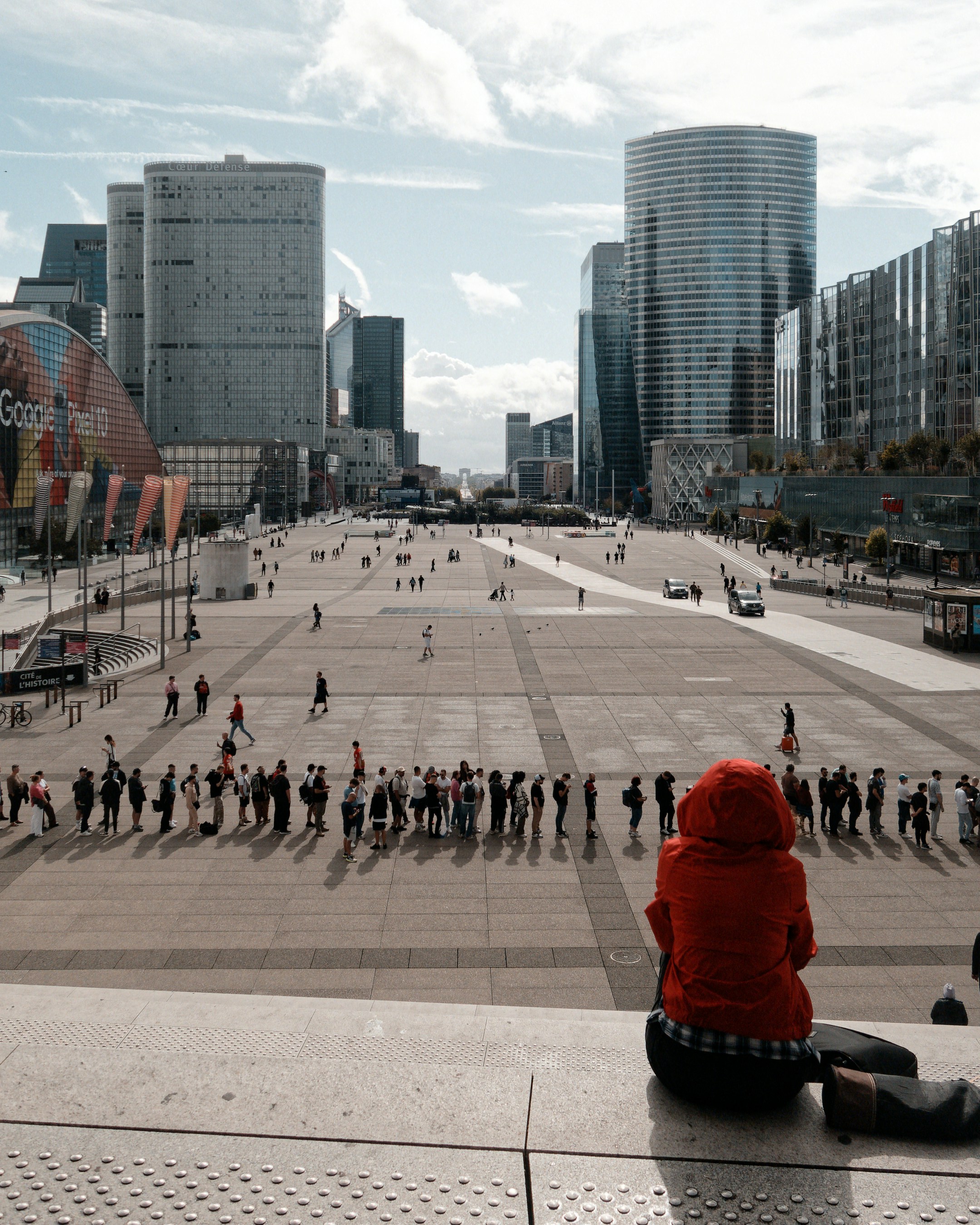 Person in red hoodie overlooks bustling urban plaza with skyscrapers