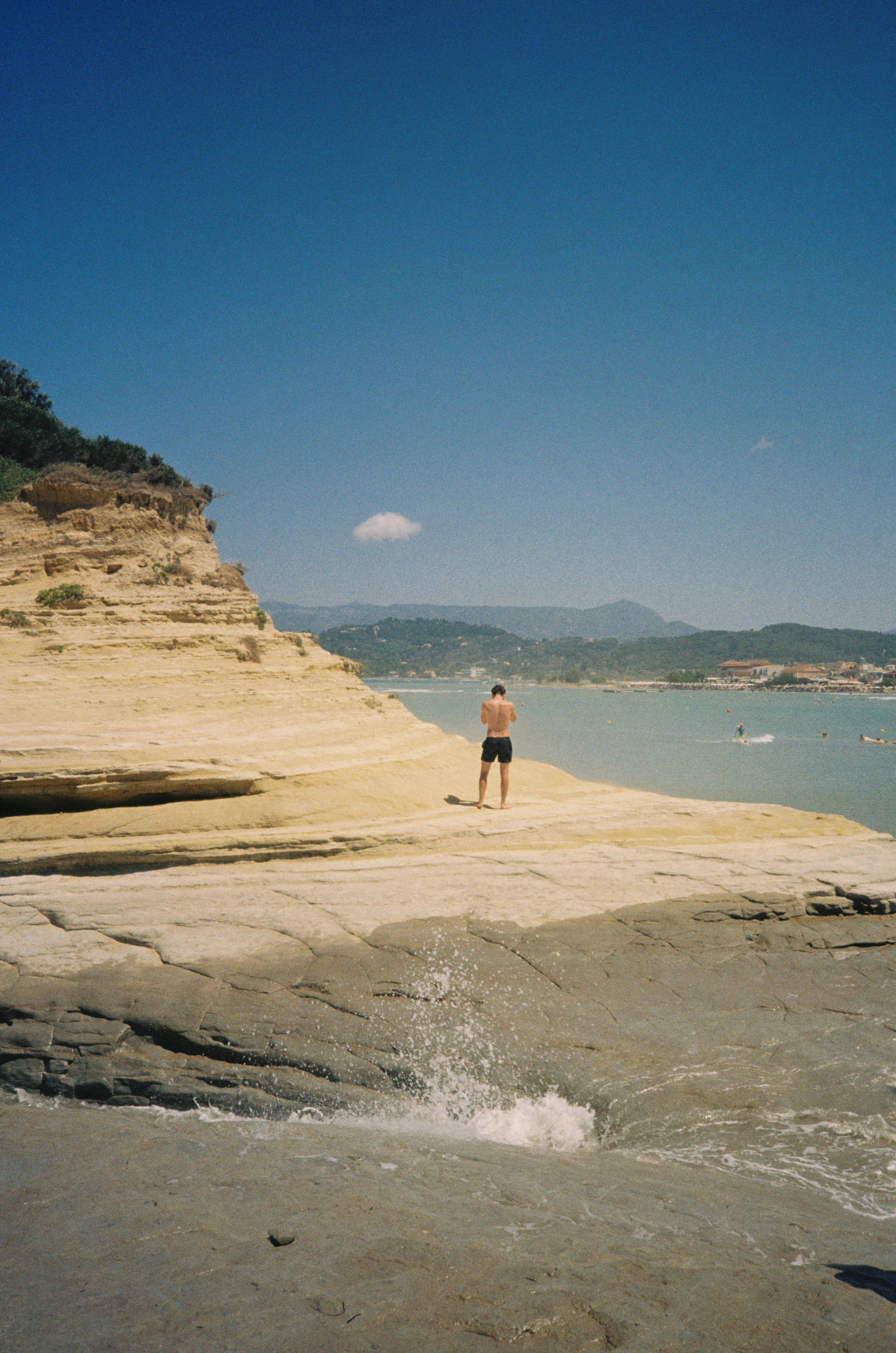 Man stands on rocky beach by cliffs and blue sea.