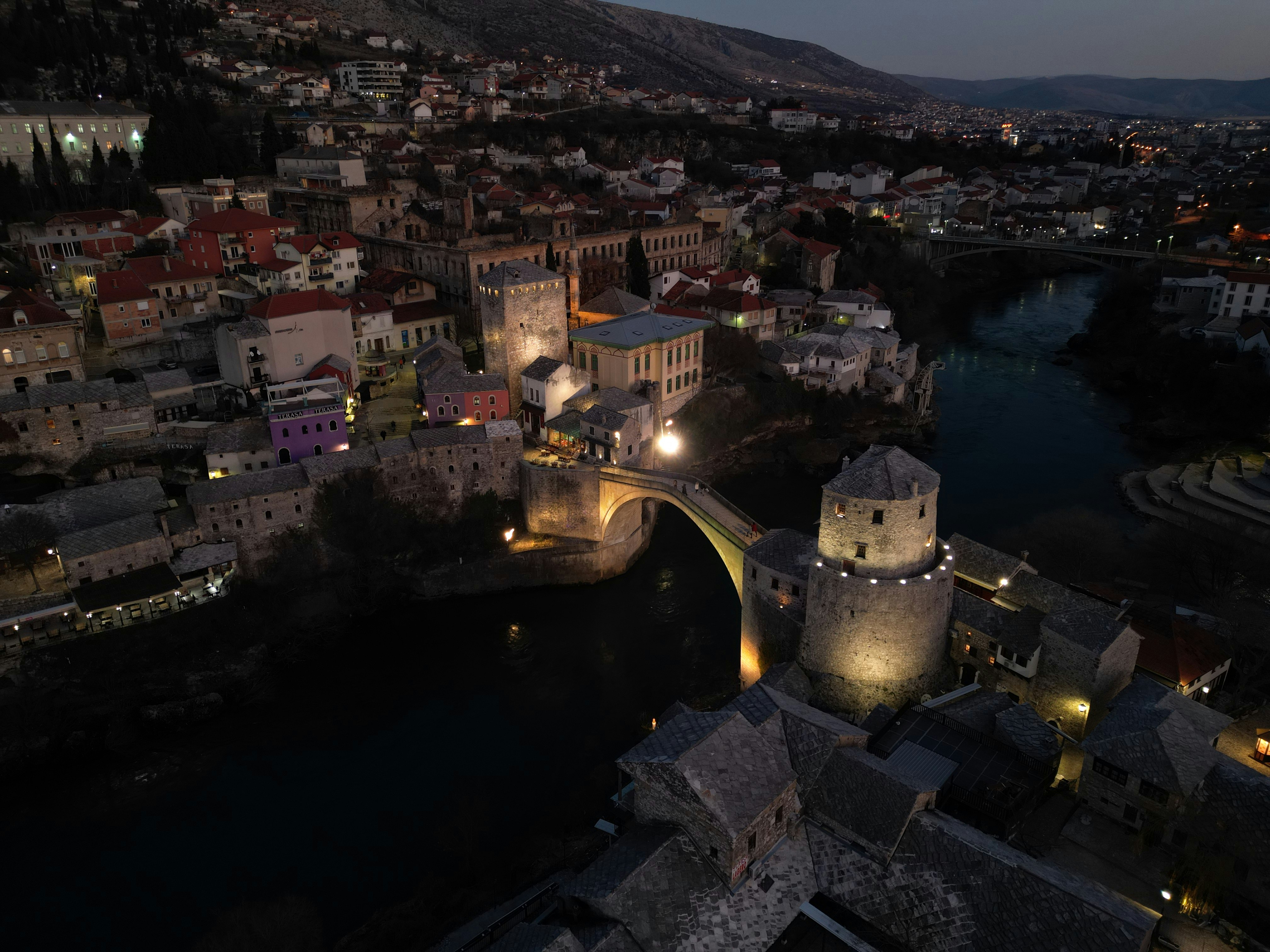 Stari most bridge and city illuminated at night with mountains.