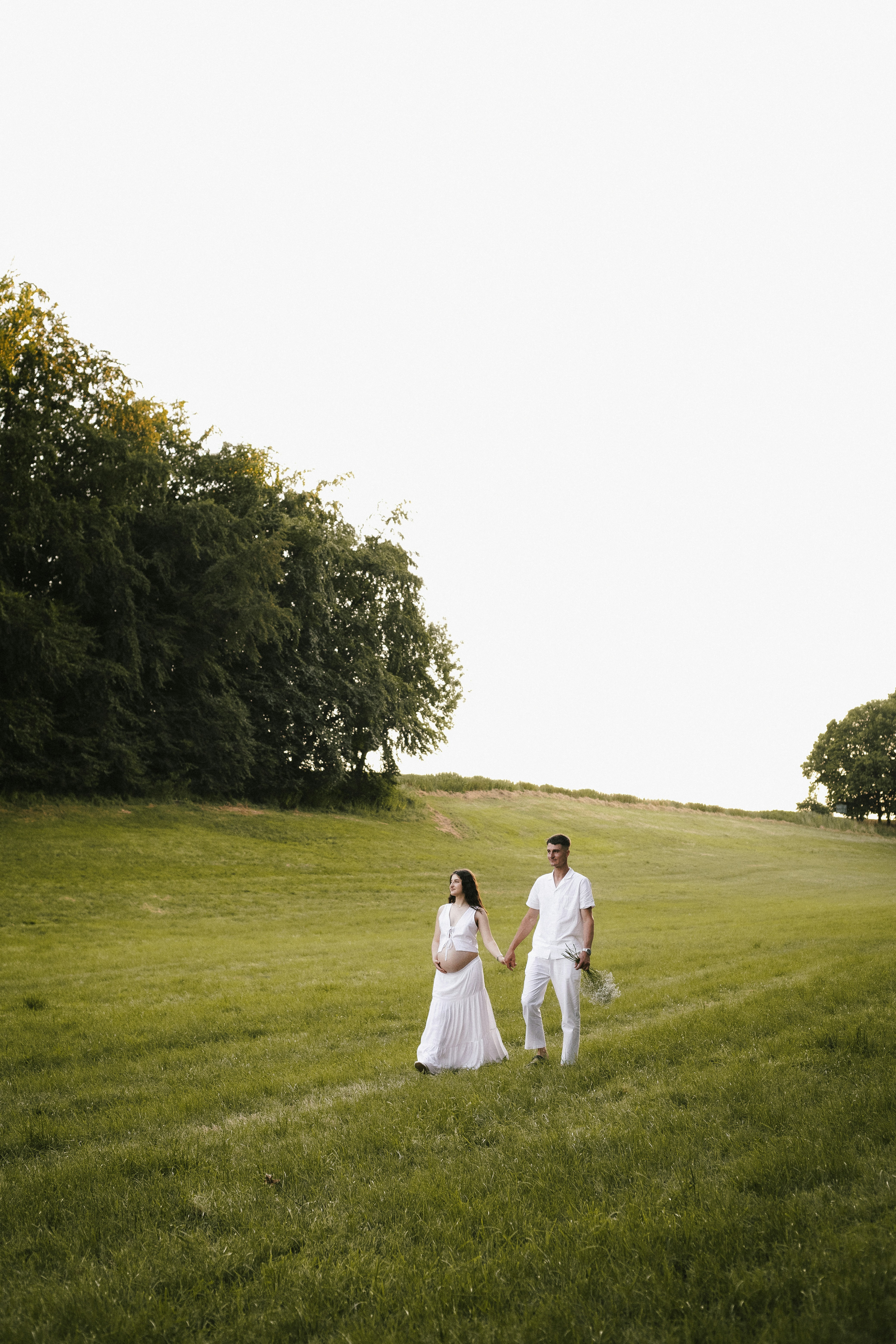 A pregnant couple walks hand-in-hand through a green field.
