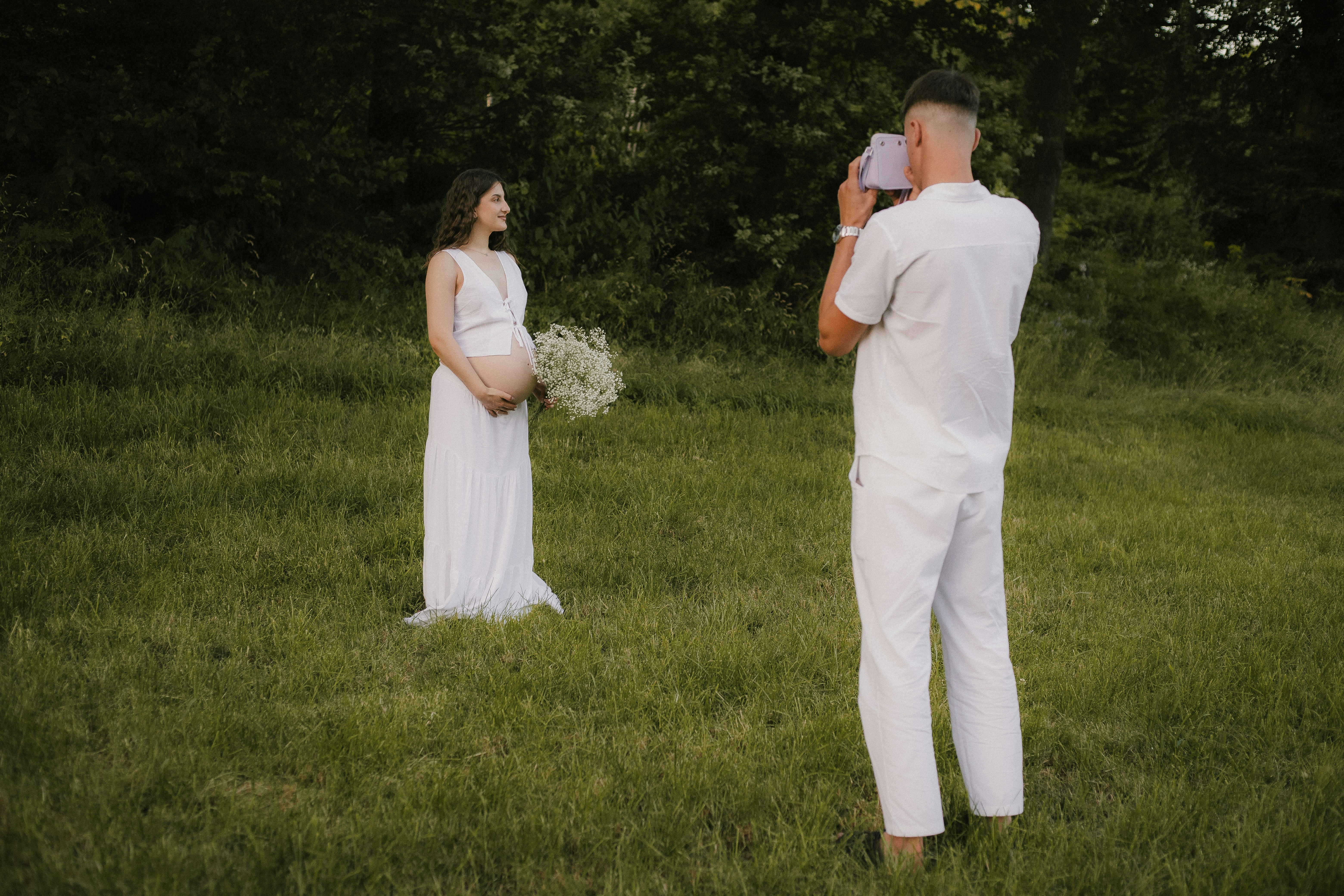 Man photographs pregnant woman holding flowers in a field.