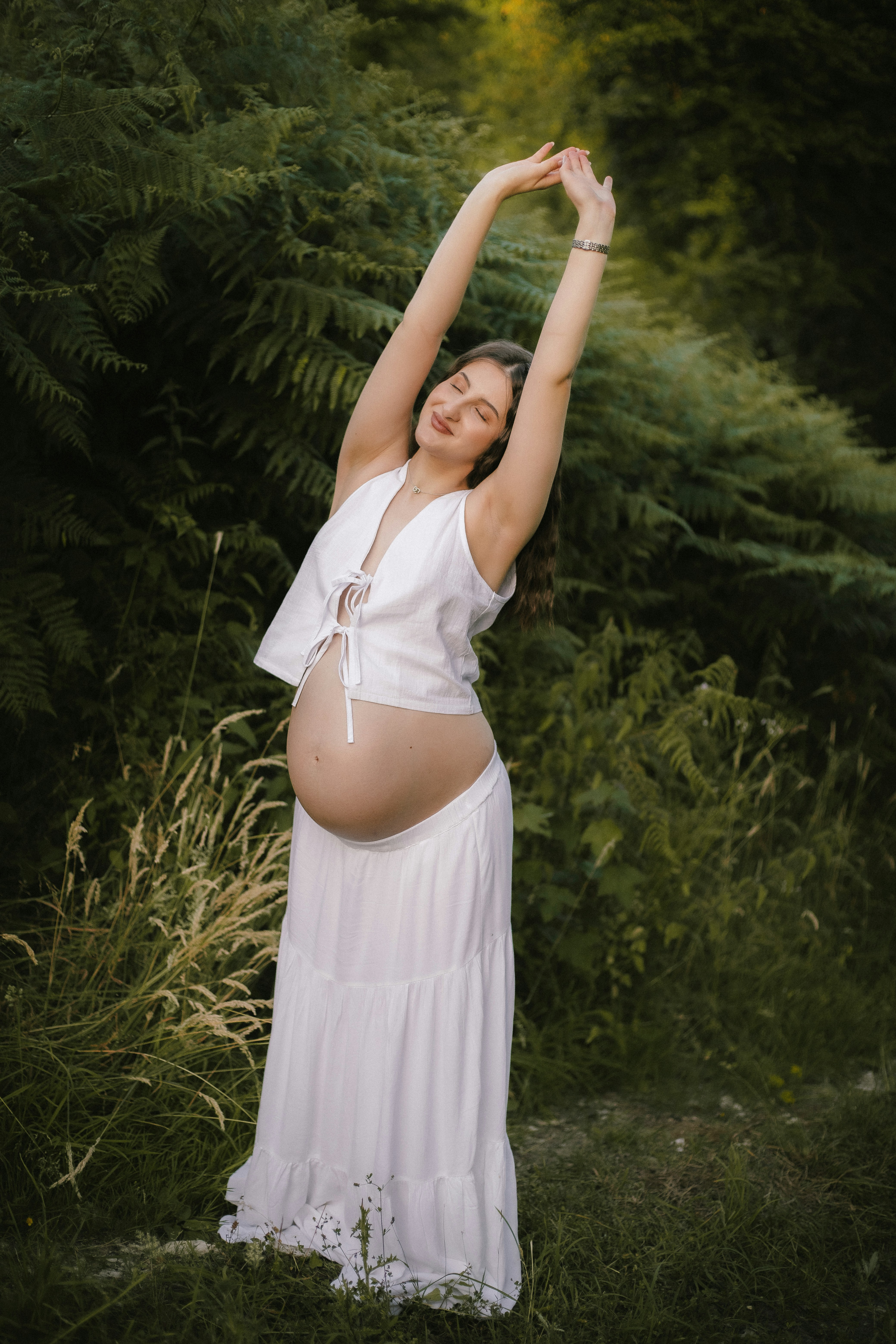 Pregnant woman in white stretching outdoors among green foliage.